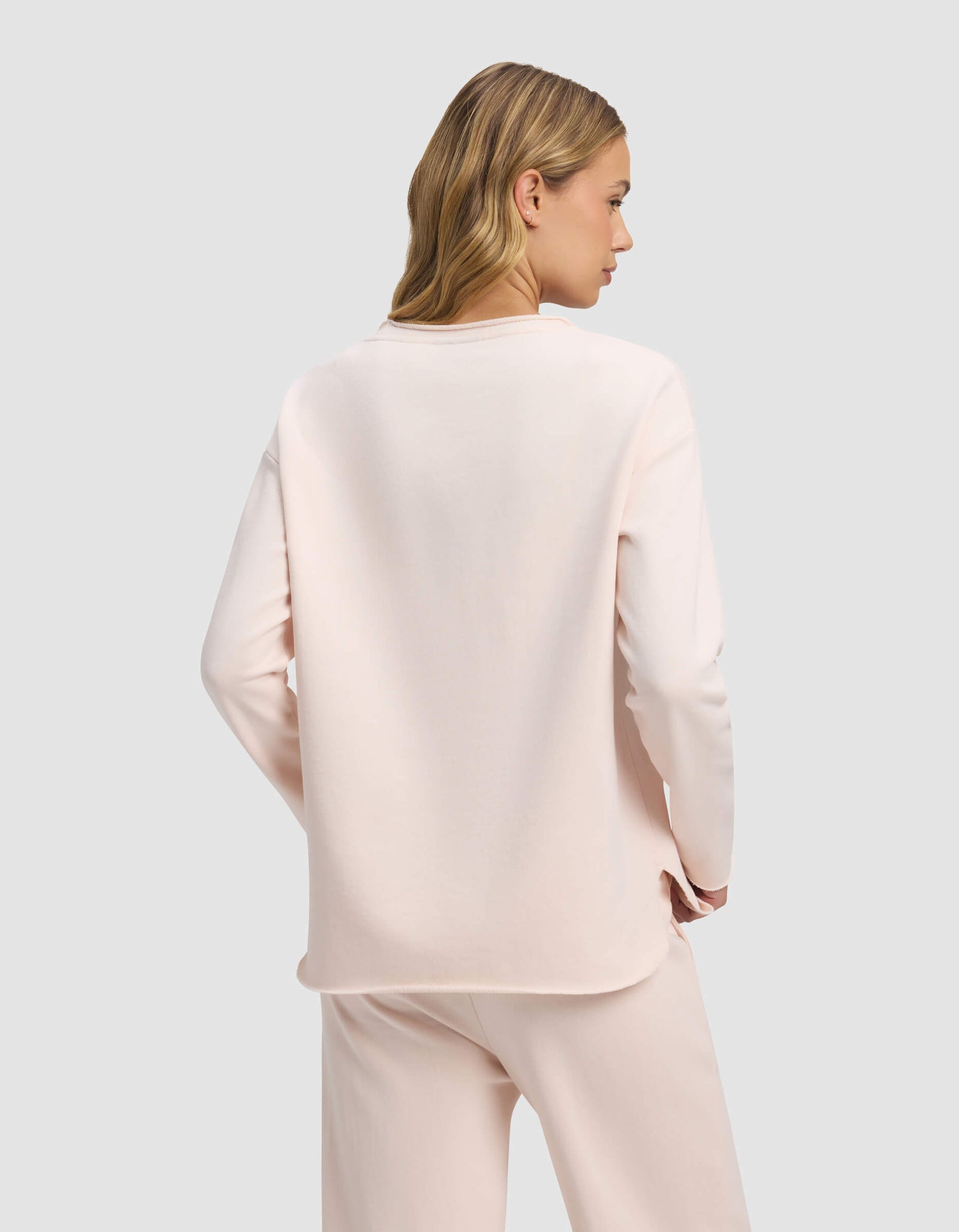 A woman with blonde hair models the Cozy Earth Women's Chelsea Pullover in light pink, standing with her back to the camera against a plain light grey background.