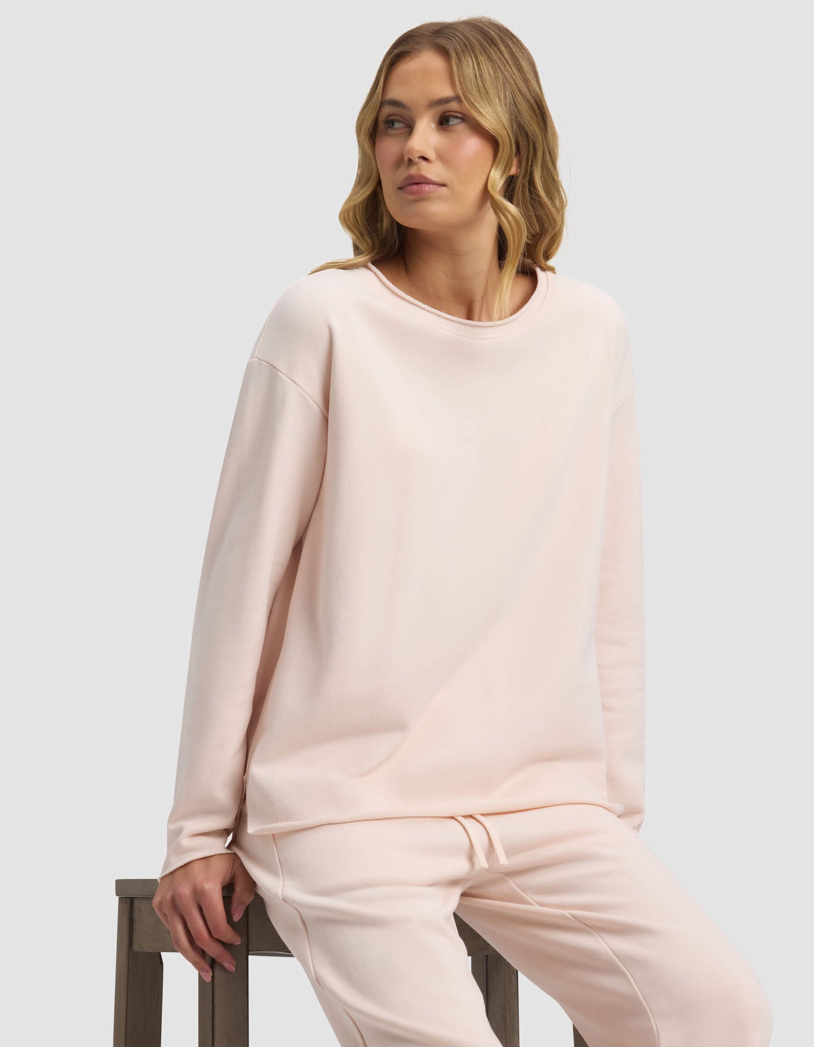 A woman with wavy blonde hair sits on a wooden stool, wearing the Cozy Earth Women's Chelsea Pullover in light pink. She gazes to her left with a neutral expression against a plain light background.