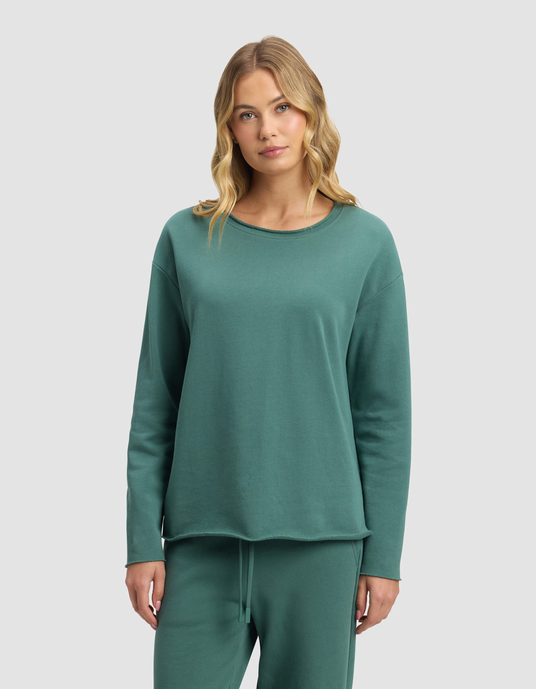 A woman with blonde wavy hair wears the Cozy Earth Women's Chelsea Pullover in green with matching pants, standing facing forward against a plain light gray background.