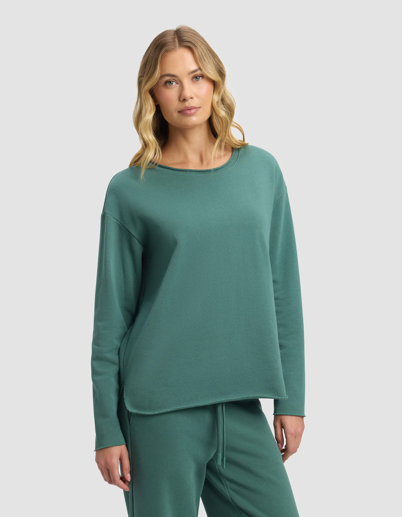 A woman with wavy blonde hair models the Cozy Earth Women's Chelsea Pullover and matching pants in green, standing against a plain light gray background.