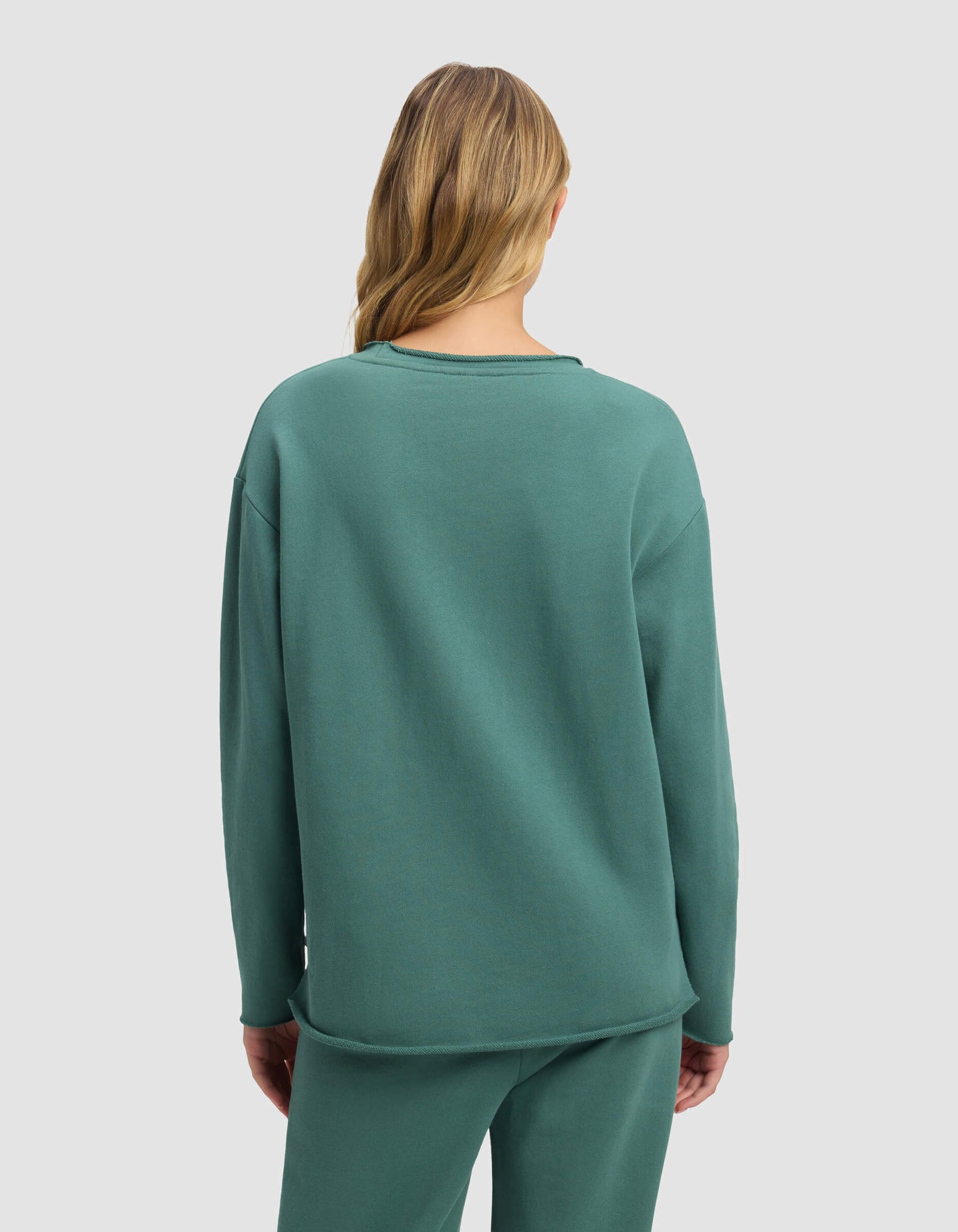 A woman with long, wavy blonde hair is seen from behind wearing the Cozy Earth Women's Chelsea Pullover in teal, paired with matching pants, set against a plain light gray background.