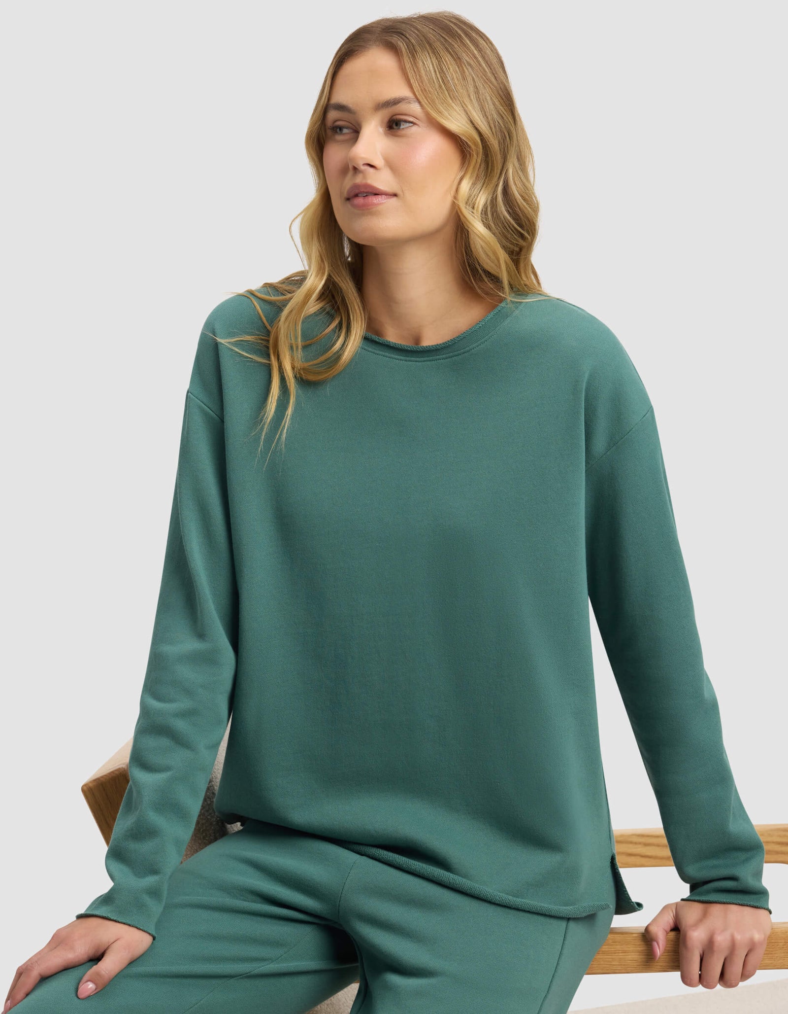 A woman with long blonde hair sits on a wooden bench wearing the Cozy Earth Women's Chelsea Pullover in teal, paired with matching pants, gazing slightly to the side against a plain background.