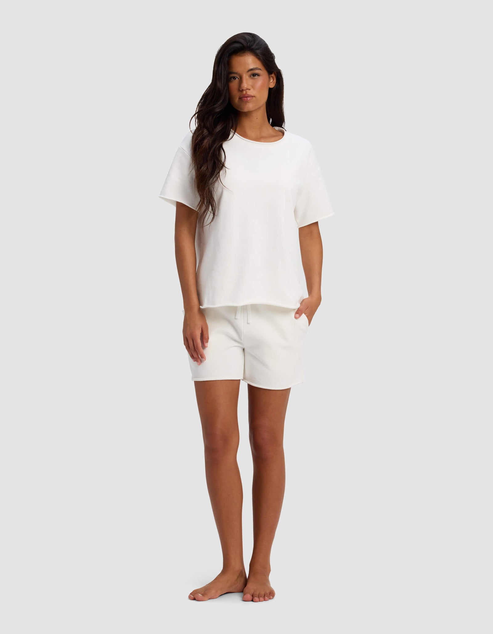 A woman with long dark hair stands barefoot against a light gray background, wearing the Cozy Earth Women's Chelsea Short Sleeve Set—loose-fitting white T-shirt and matching shorts, with her left hand in her pocket.