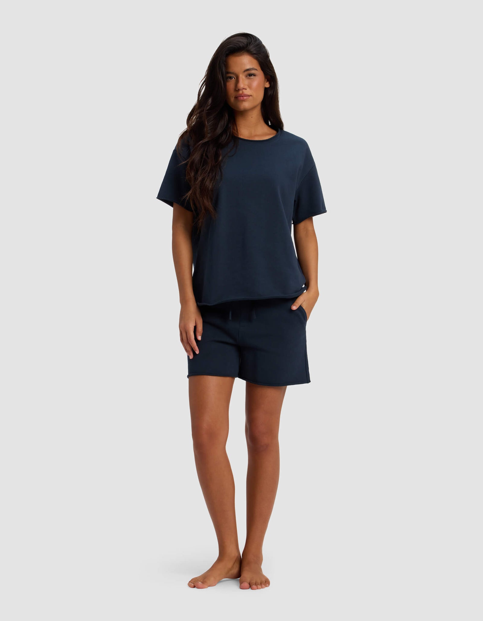 A woman with long dark hair stands barefoot against a plain light background, wearing the Cozy Earth Women's Chelsea Short Sleeve Set in dark navy, with her hands in her pockets.