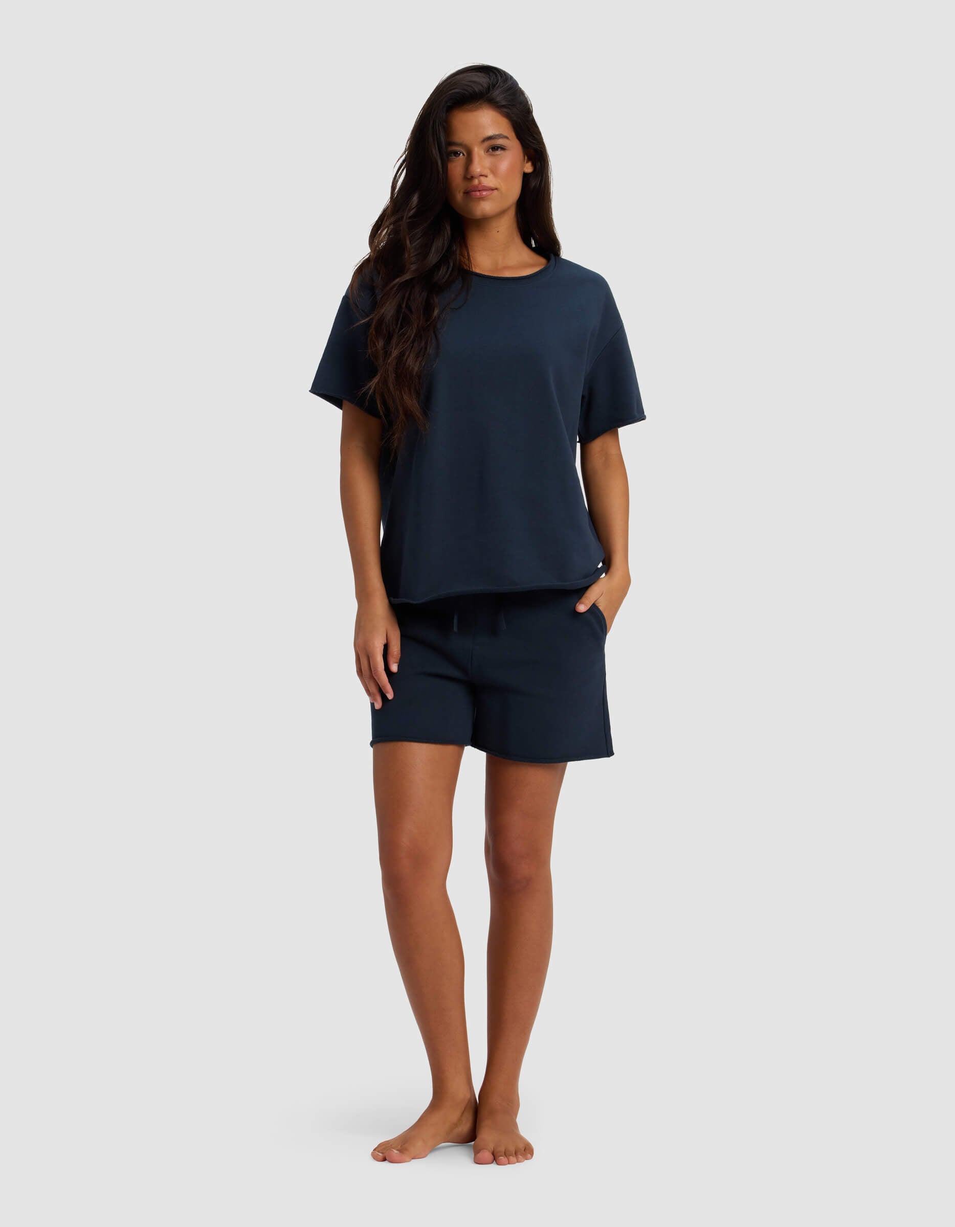 A woman with long dark hair stands barefoot against a plain light background, wearing the Cozy Earth Women's Chelsea Short Sleeve Set in dark navy, with her hands in her pockets.