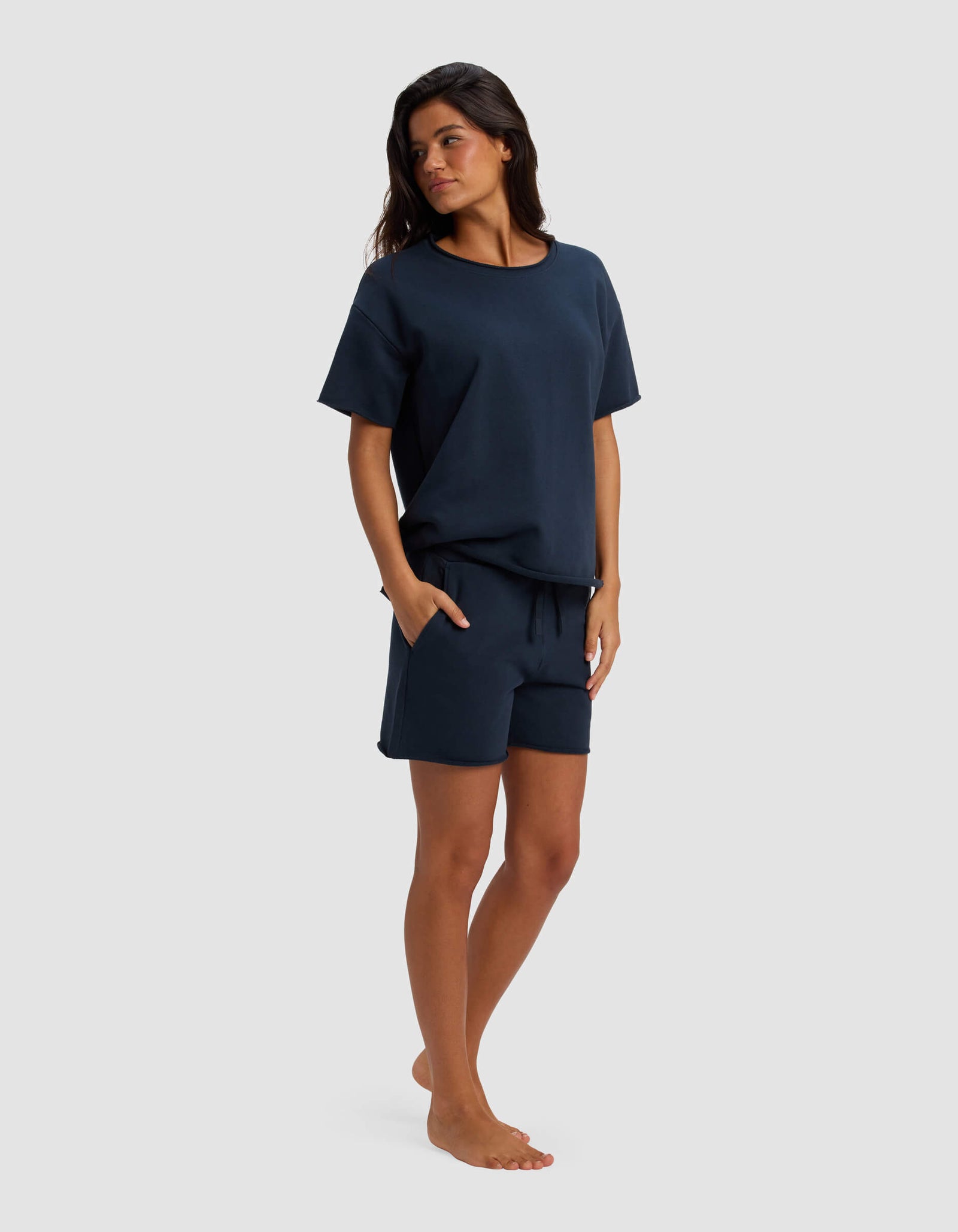 A woman stands barefoot against a light gray background, wearing the Cozy Earth Women's Chelsea Short Sleeve Set in navy blue. The relaxed-fit top and matching shorts feature one hand in the pocket as she gazes off to the side.