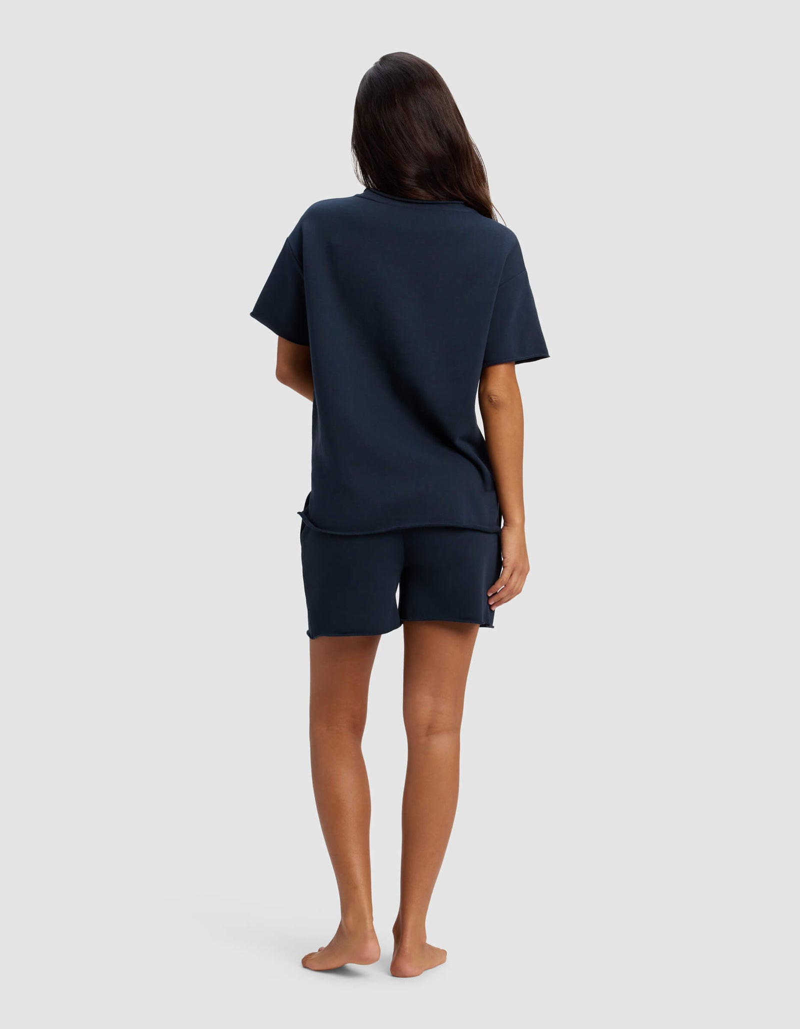 A person with long dark hair stands barefoot, facing away, wearing the Cozy Earth Women's Chelsea Short Sleeve Set in navy against a plain light grey background.