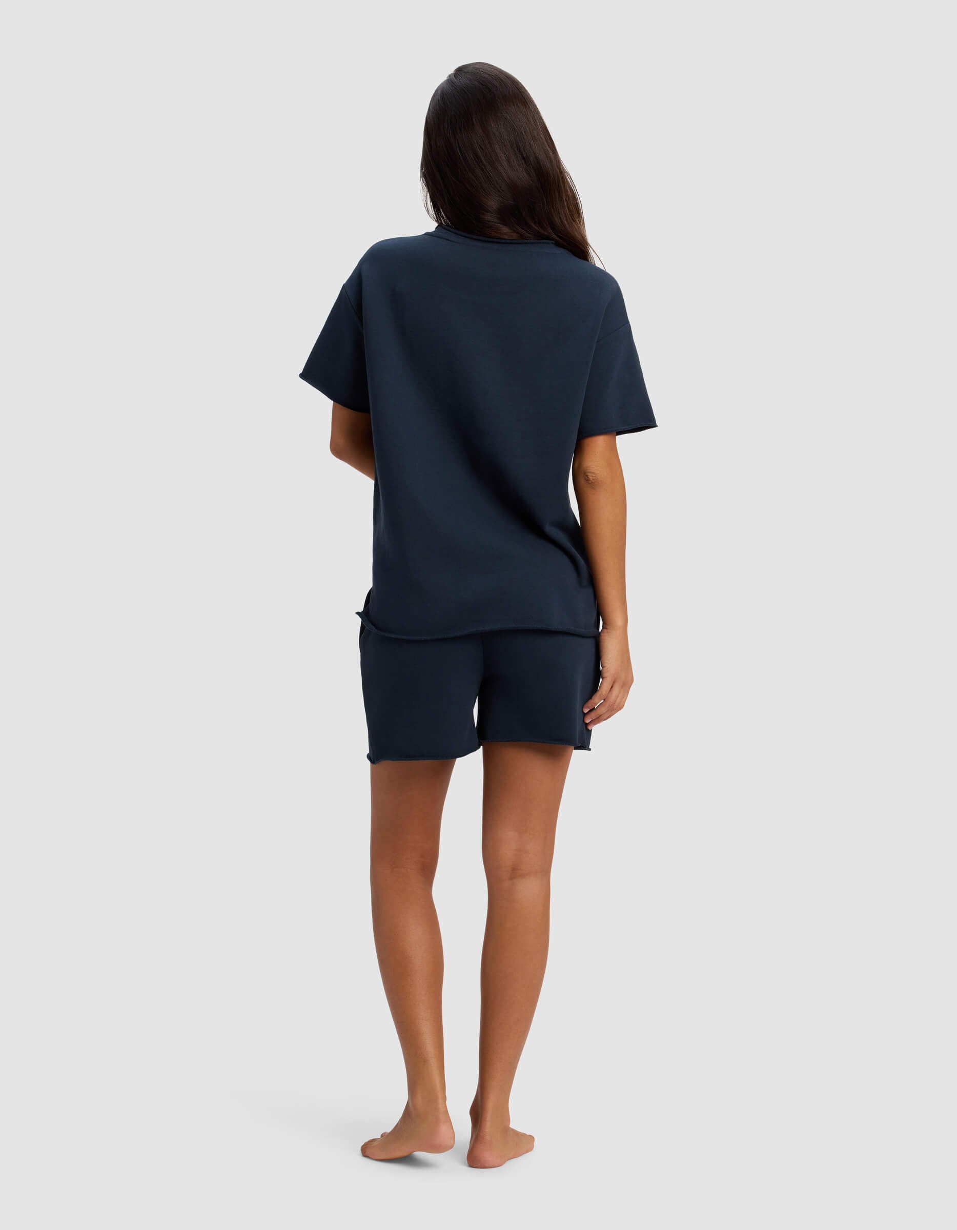 A person with long dark hair stands barefoot, facing away, wearing the Cozy Earth Women's Chelsea Short Sleeve Set in navy against a plain light grey background.