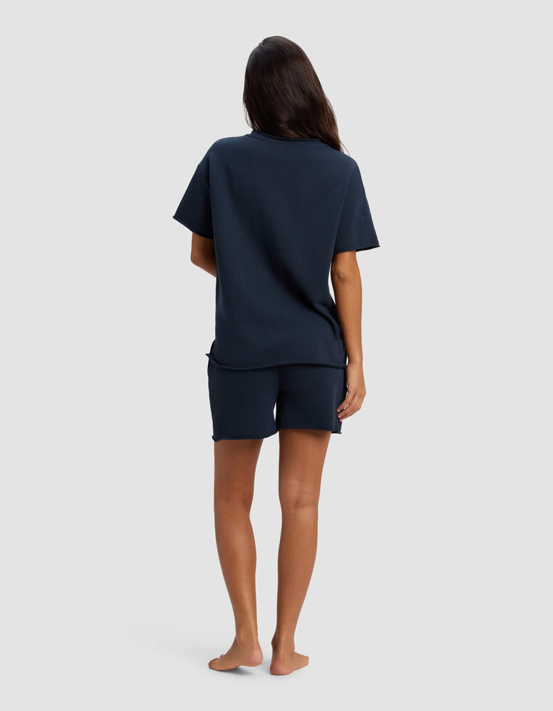 A person with long dark hair stands barefoot, facing away, wearing the Cozy Earth Women's Chelsea Short Sleeve Set in navy against a plain light grey background.