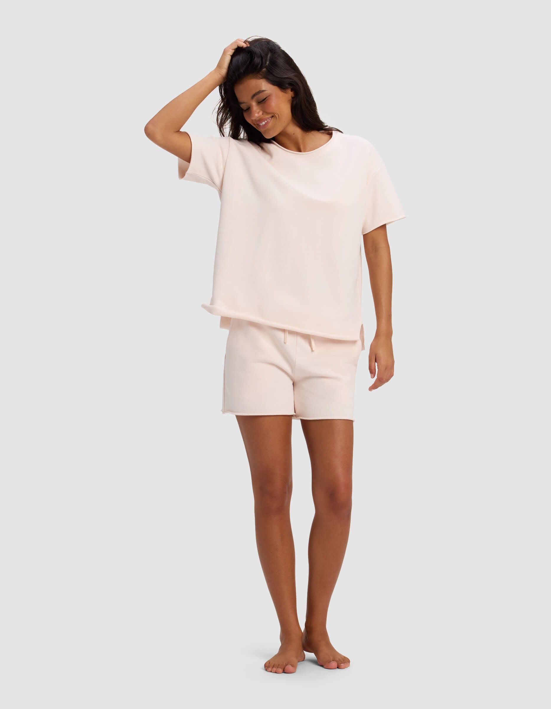 A woman smiles barefoot against a gray background, wearing the Cozy Earth Women's Chelsea Short Sleeve Set in light pink. One hand touches her head while the other rests by her side.