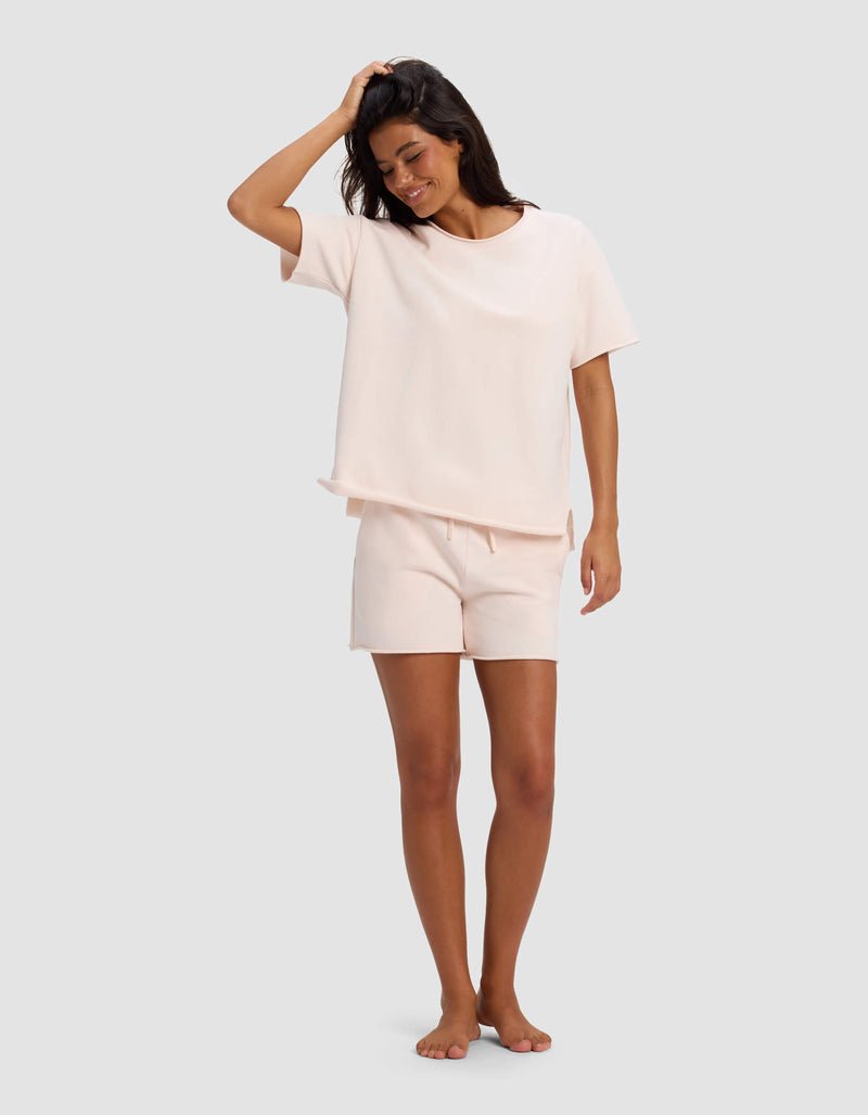 A woman smiles barefoot against a gray background, wearing the Cozy Earth Women's Chelsea Short Sleeve Set in light pink. One hand touches her head while the other rests by her side.