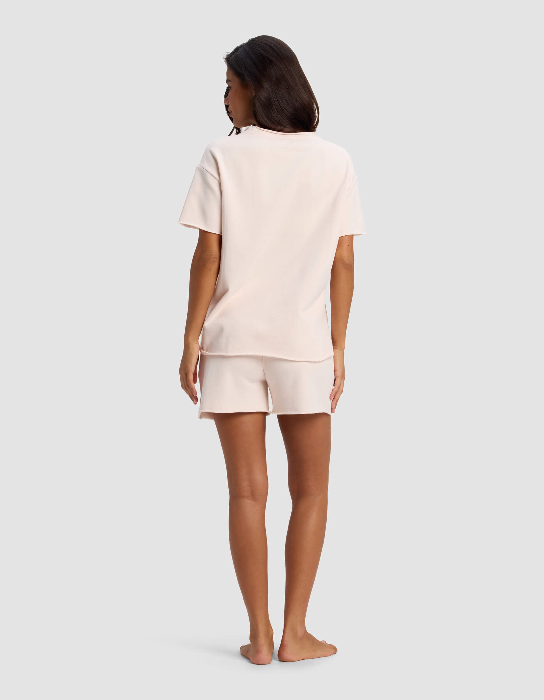 A woman stands barefoot facing away, wearing the Cozy Earth Women's Chelsea Short Sleeve Set in light pink, against a plain light gray background.