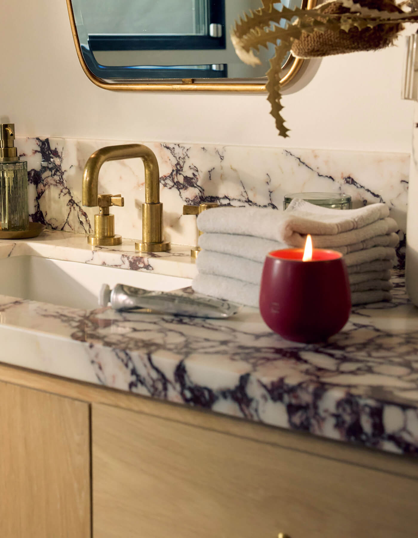 A marble bathroom countertop holds a Cozy Earth Candle CIDER, a gold faucet, folded beige towels, a glass soap dispenser, and a mirror with a gold frame in the background.