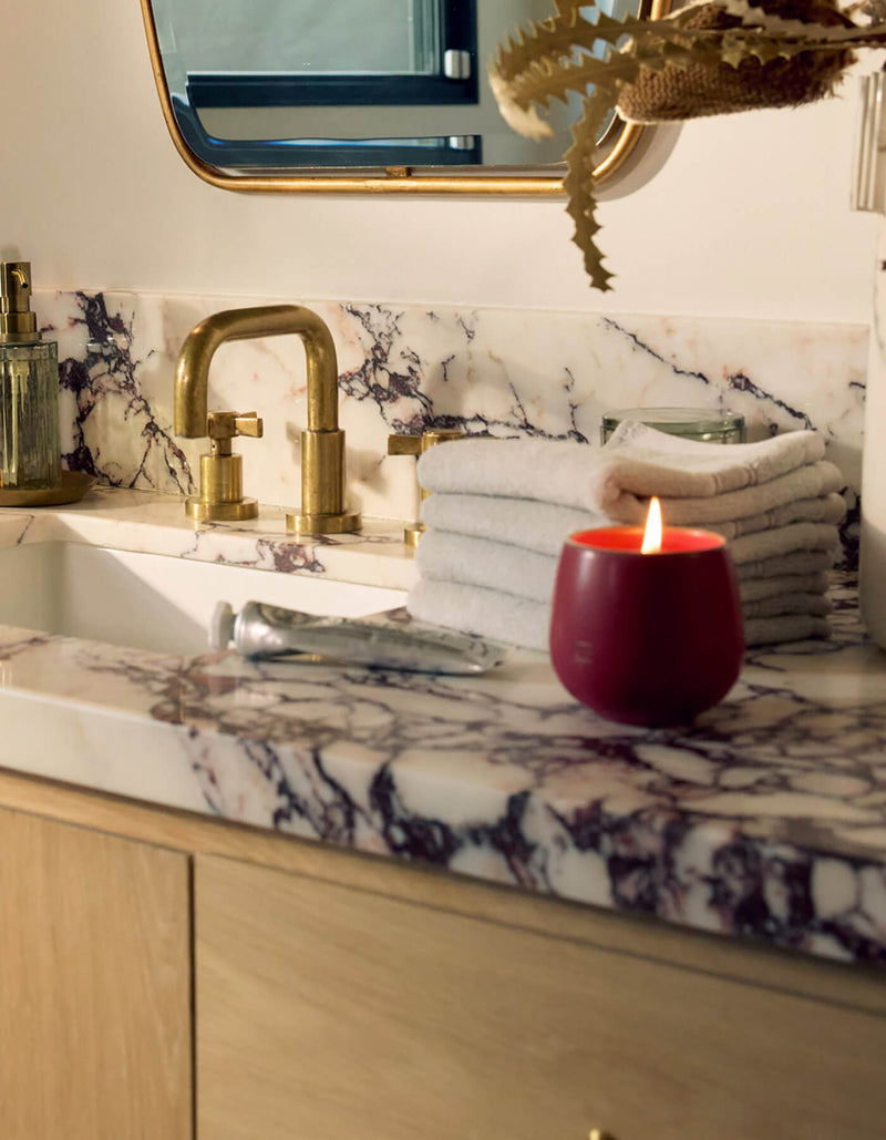 A marble bathroom countertop holds a Cozy Earth Candle CIDER, a gold faucet, folded beige towels, a glass soap dispenser, and a mirror with a gold frame in the background.