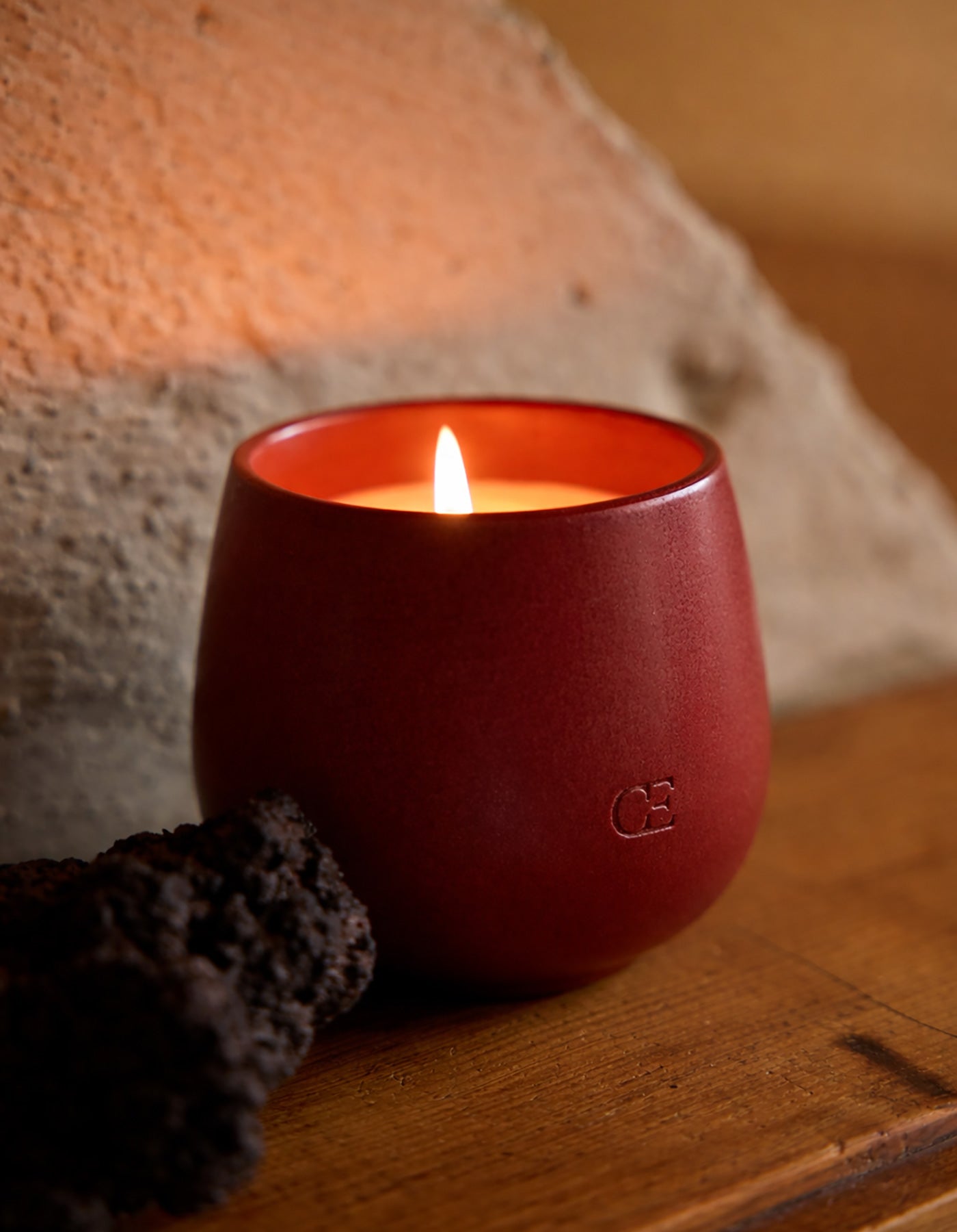 The Cozy Earth Candle CIDER, in a round matte red holder, sits on a wooden surface beside dark porous rock with a textured stone wall behind it.