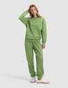 A woman in the Cozy Earth Women's Cityscape 2.0 Crewneck & Sweatpant Set, light green, stands casually with one hand touching her head. She pairs the outfit with white sneakers and looks slightly to the side against a plain background.|Color:Clover