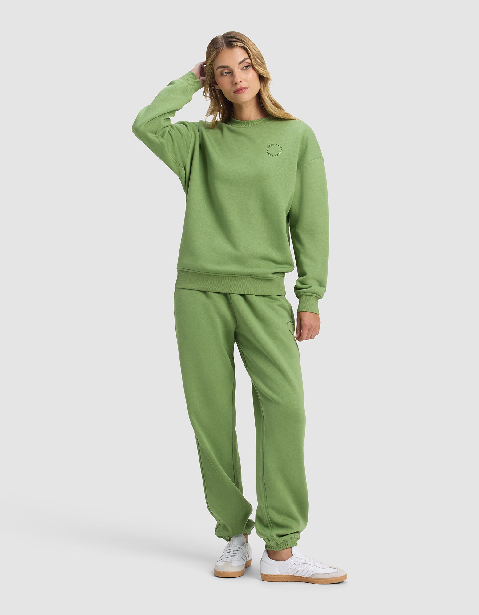 A woman in the Cozy Earth Women's Cityscape 2.0 Crewneck & Sweatpant Set, light green, stands casually with one hand touching her head. She pairs the outfit with white sneakers and looks slightly to the side against a plain background.|Color:Clover