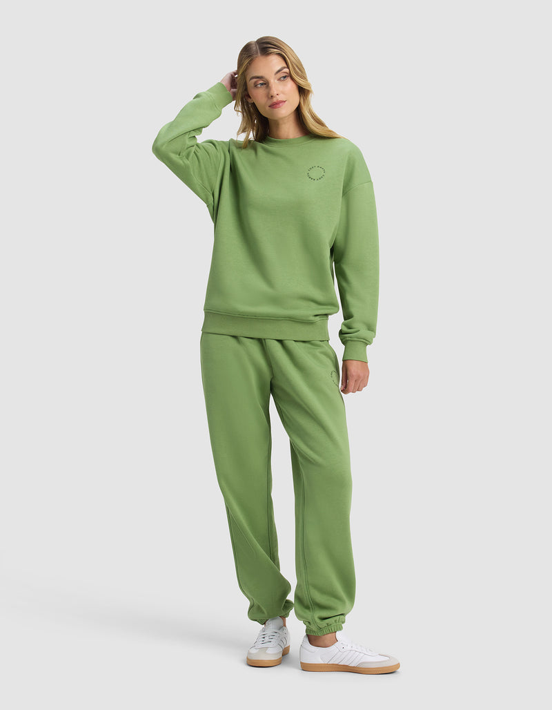 A woman in the Cozy Earth Women's Cityscape 2.0 Crewneck & Sweatpant Set, light green, stands casually with one hand touching her head. She pairs the outfit with white sneakers and looks slightly to the side against a plain background.