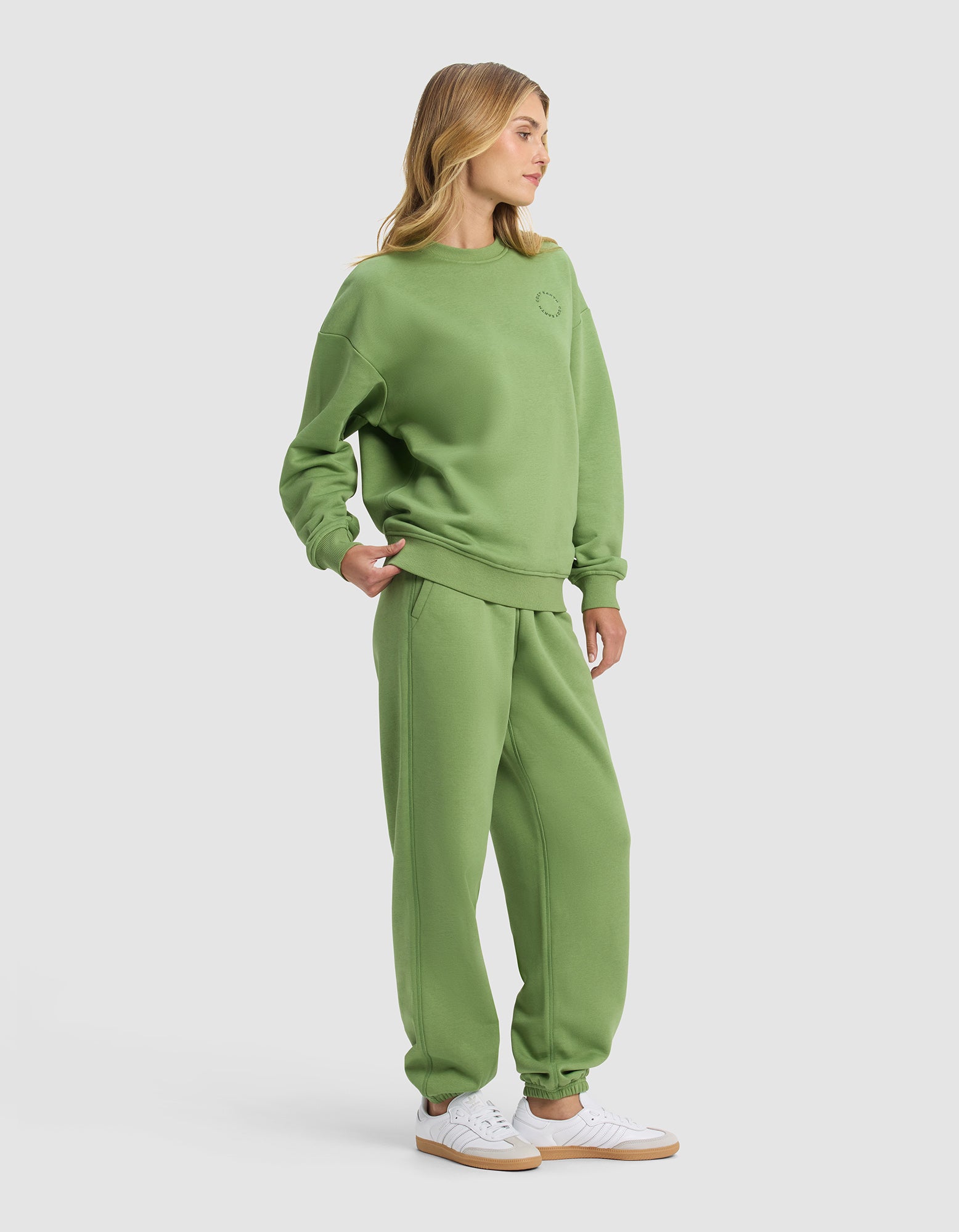 A woman models the Cozy Earth Women's Cityscape 2.0 Crewneck & Sweatpant Set in green with white sneakers against a plain light gray background, standing slightly to the side.|Color:Clover
