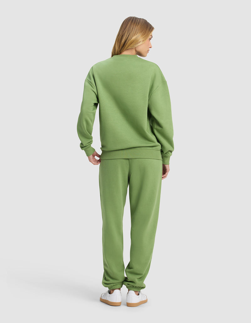 A person with long blonde hair faces away, wearing Cozy Earth's Women's Cityscape 2.0 Crewneck & Sweatpant Set in light green with white sneakers, set against a plain light gray background.