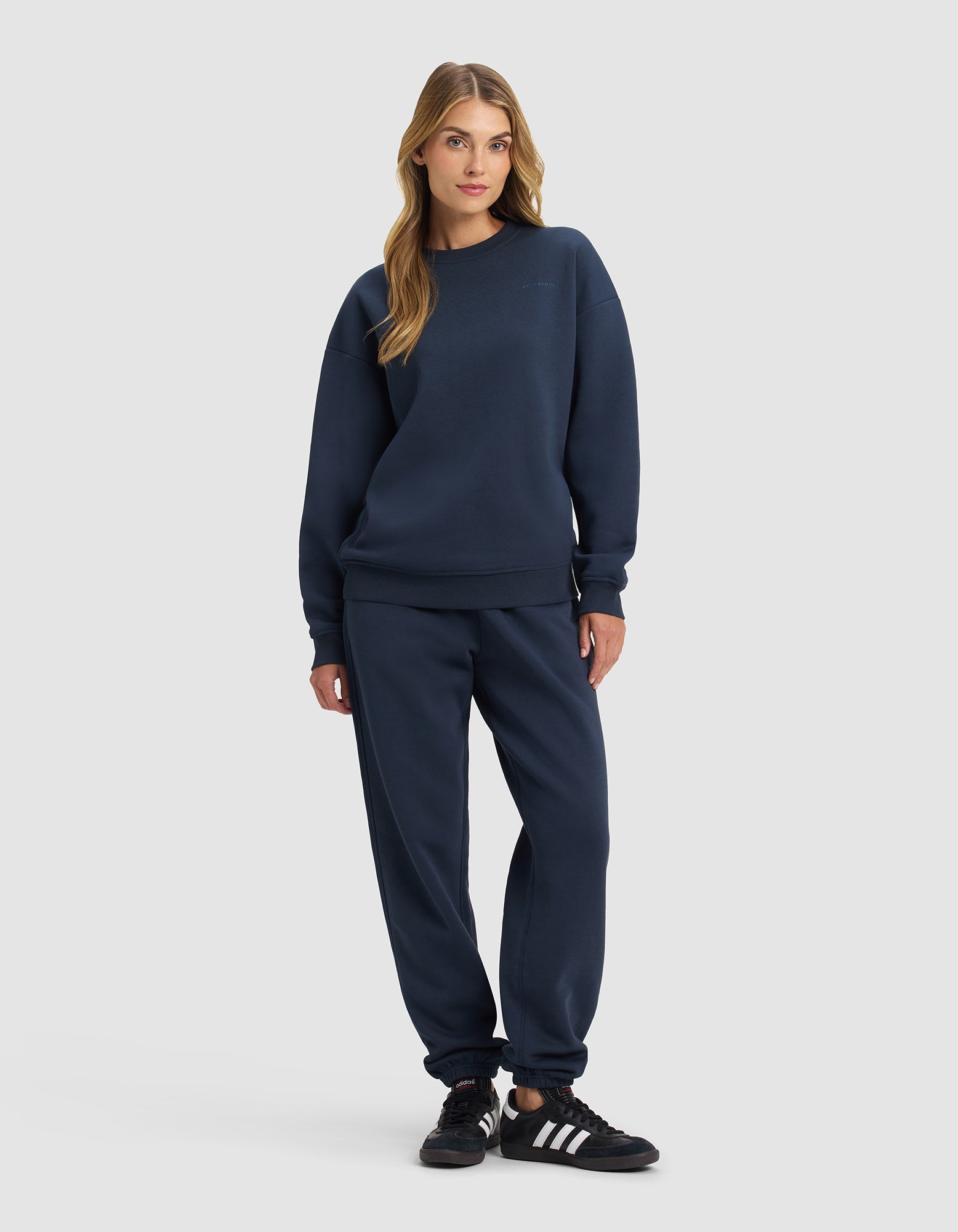 A woman faces forward in the Cozy Earth Women's Cityscape 2.0 Crewneck & Sweatpant Set, navy blue, paired with black and white athletic shoes against a plain light background.|Color:Eclipse