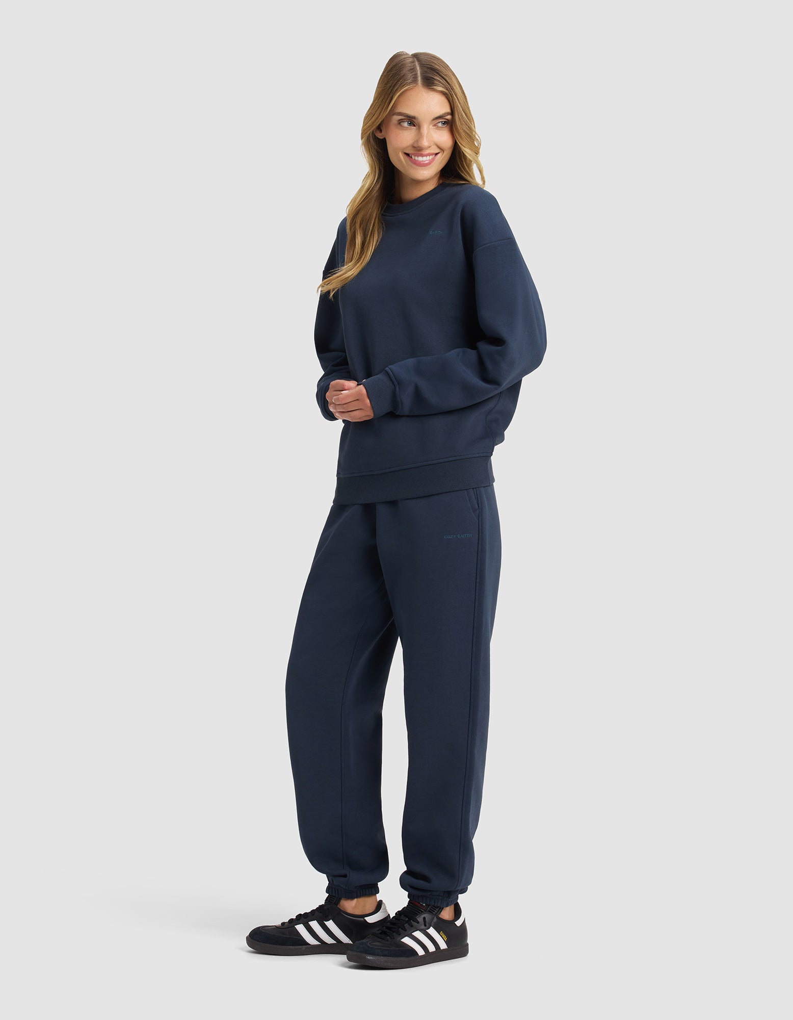 A woman with long blonde hair smiles with hands clasped, wearing the Women's Cityscape 2.0 Crewneck & Sweatpant Set from Cozy Earth and black Adidas sneakers, standing against a plain light background.
