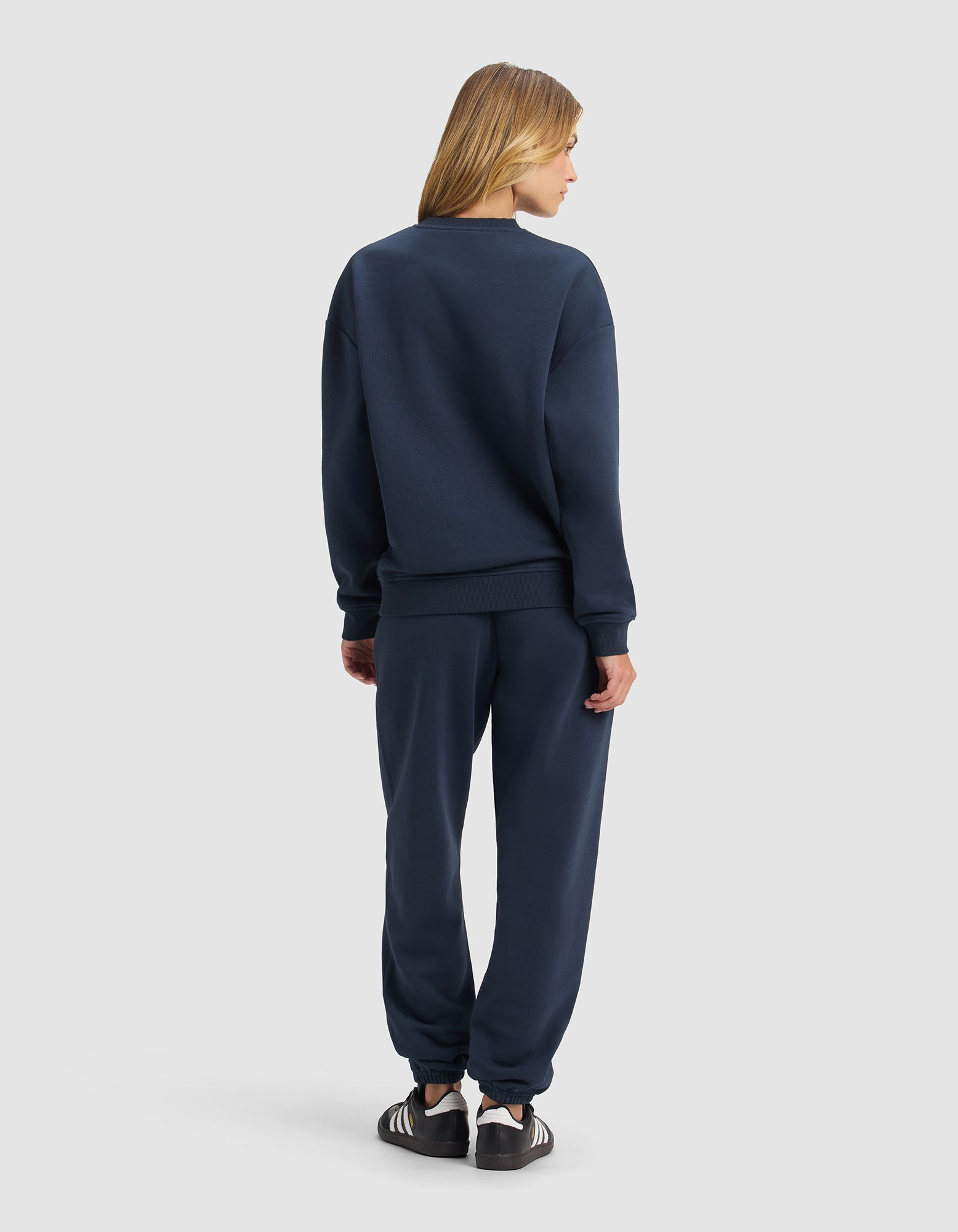 A person faces away, wearing the Cozy Earth Women's Cityscape 2.0 Crewneck & Sweatpant Set in navy blue, paired with black and white sneakers, against a plain light gray background.