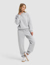 A woman models Cozy Earth’s Women's Cityscape 2.0 Crewneck & Sweatpant Set in light gray, paired with white sneakers, standing casually with one hand on her neck and gazing slightly to the side.|Color:Heather Grey