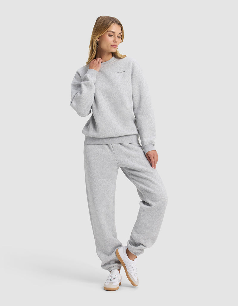 A woman models Cozy Earth’s Women's Cityscape 2.0 Crewneck & Sweatpant Set in light gray, paired with white sneakers, standing casually with one hand on her neck and gazing slightly to the side.