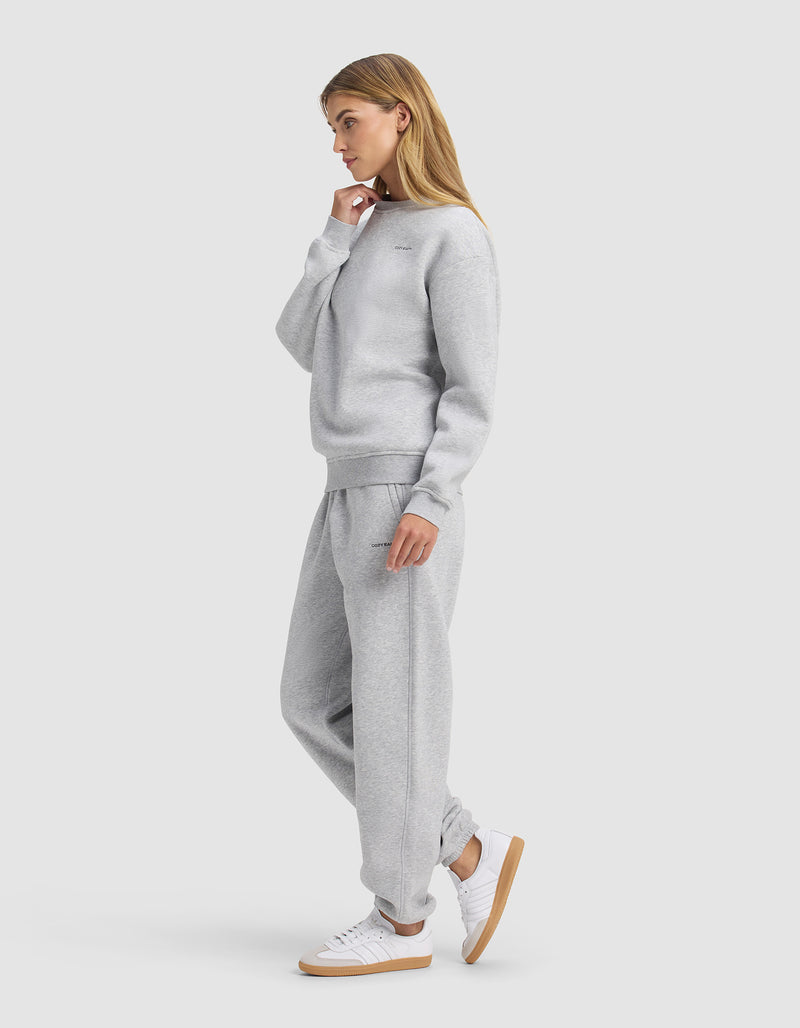 A woman in profile models the Cozy Earth Women's Cityscape 2.0 Crewneck & Sweatpant Set in light gray with white sneakers, one hand near her chin, posing against a plain light background.