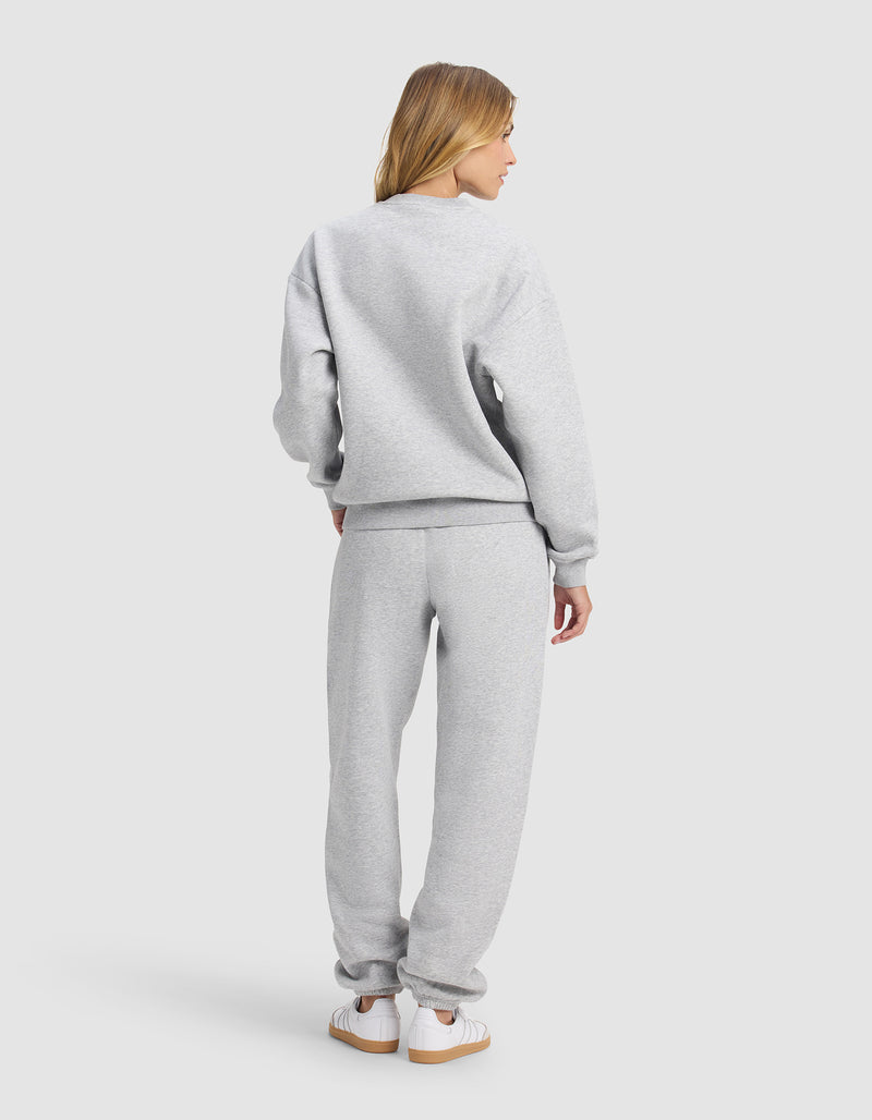 A woman faces away, wearing Cozy Earth's Women's Cityscape 2.0 Crewneck & Sweatpant Set in light gray with white sneakers, against a plain light gray background.