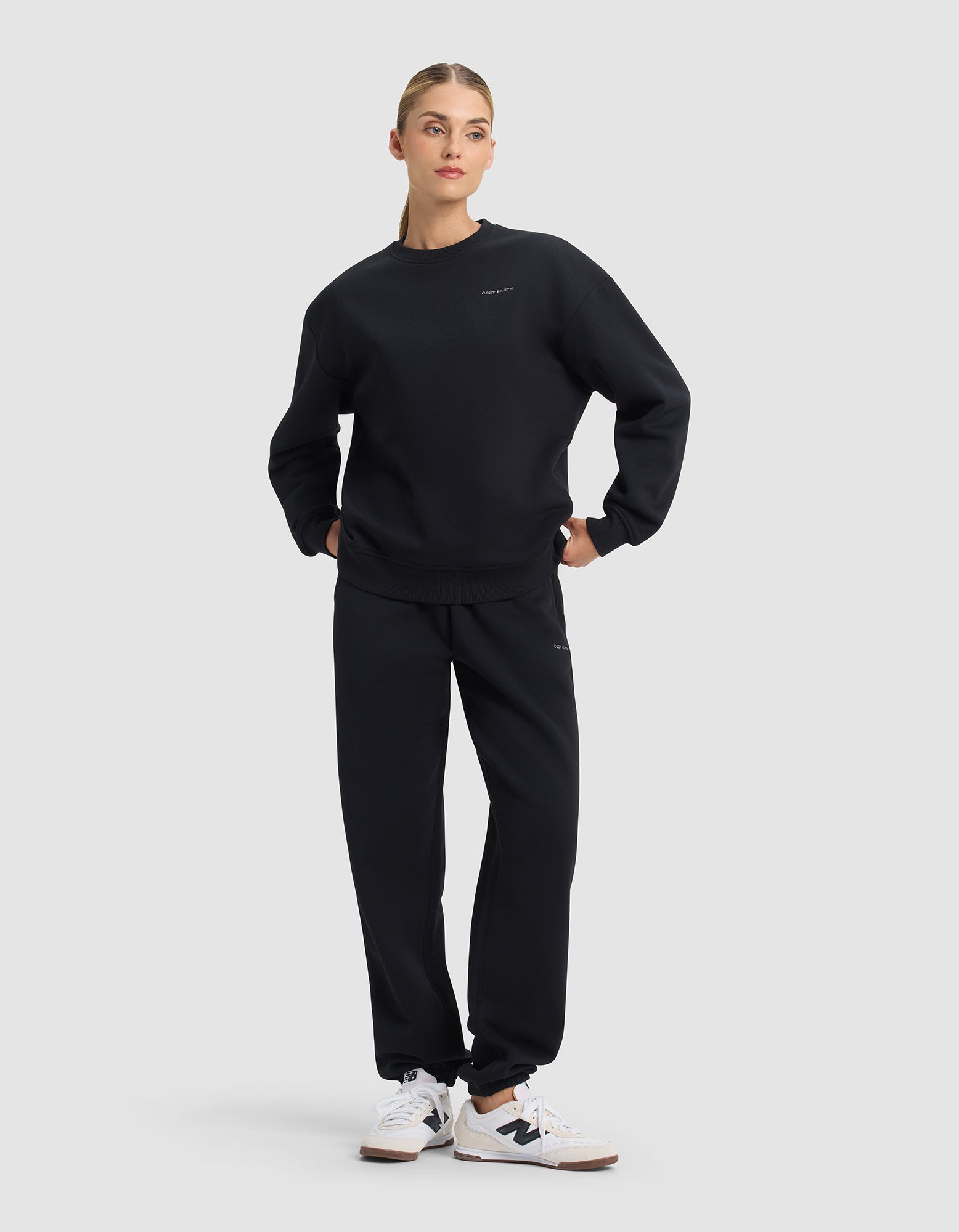 A woman wears the Cozy Earth Women's Cityscape 2.0 Crewneck & Sweatpant Set in black, paired with white sneakers. She stands against a plain background with her hands in her pockets, looking slightly to the side.|Color:Jet Black