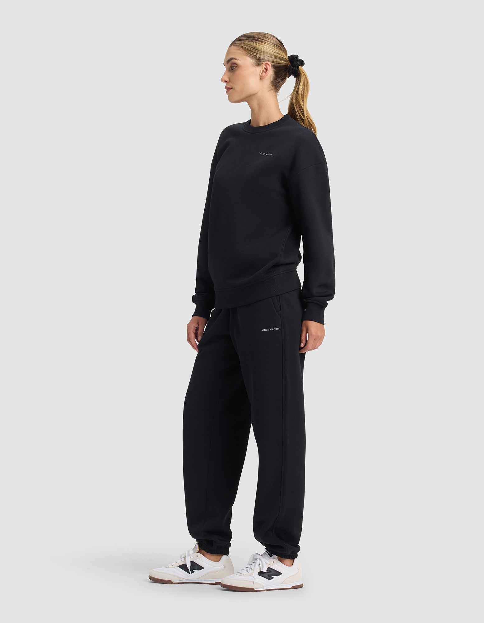 A woman stands sideways in Cozy Earth's Women's Cityscape 2.0 Crewneck & Sweatpant Set with white sneakers and a ponytail, set against a light gray background.