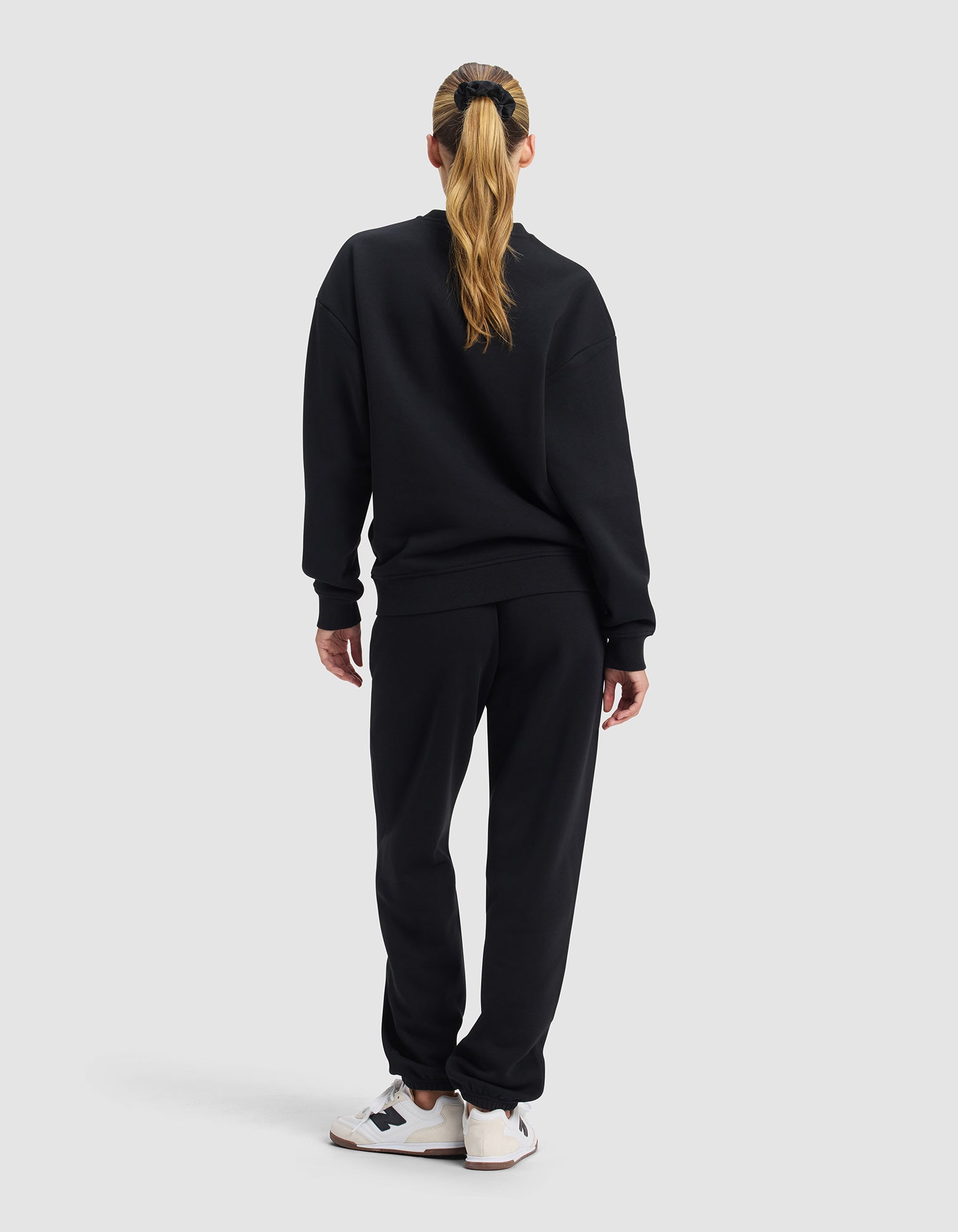 A woman with long blonde hair in a ponytail stands facing away, dressed in Cozy Earth's Women's Cityscape 2.0 Crewneck & Sweatpant Set and white sneakers with black accents, set against a plain light gray background.
