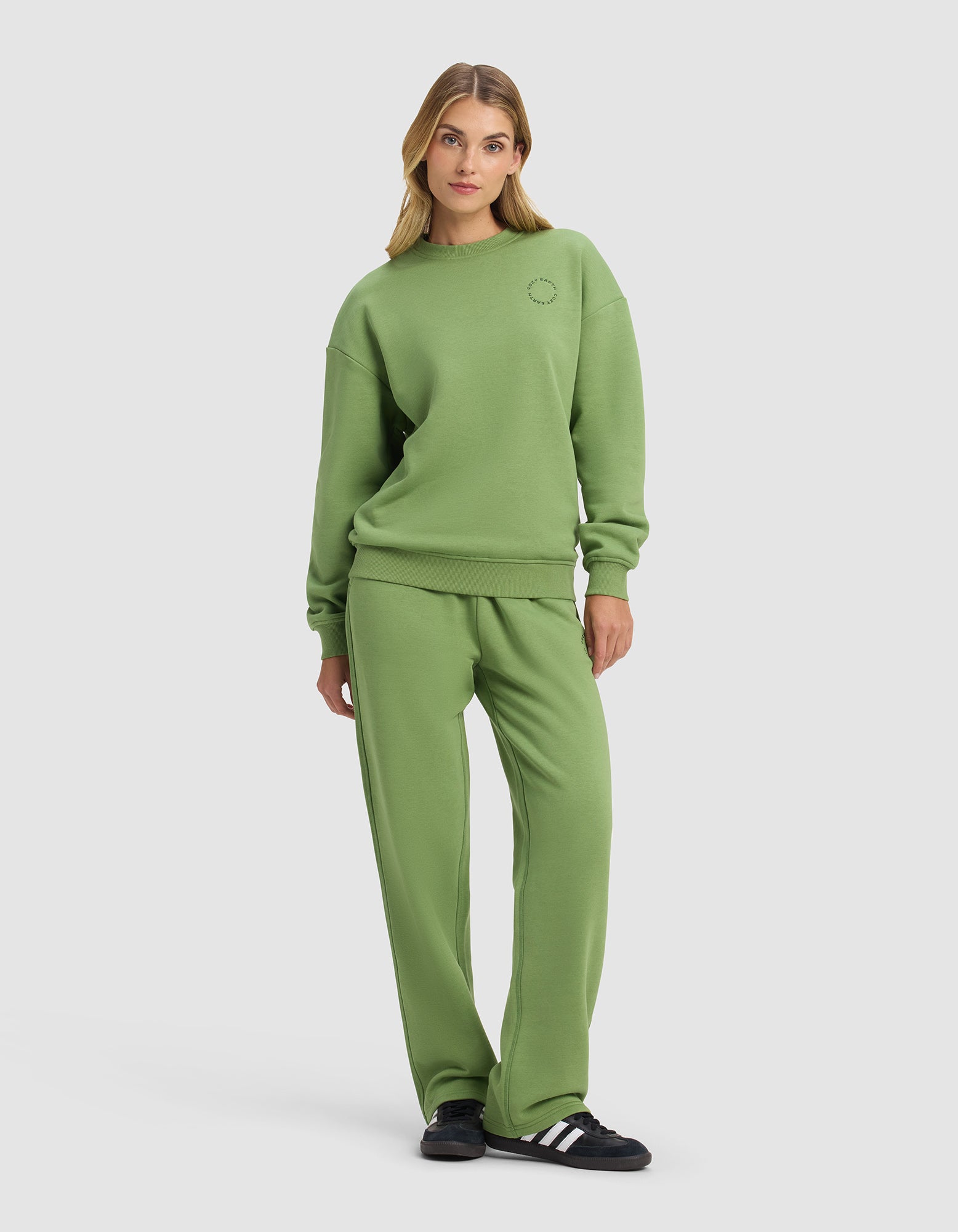 A woman poses casually in the Cozy Earth Women's Cityscape 2.0 Crewneck & Wide Leg Pant Set, featuring a relaxed fit. She stands against a plain background with one hand in her pocket, completing the look with black and white sneakers.|Color:Clover