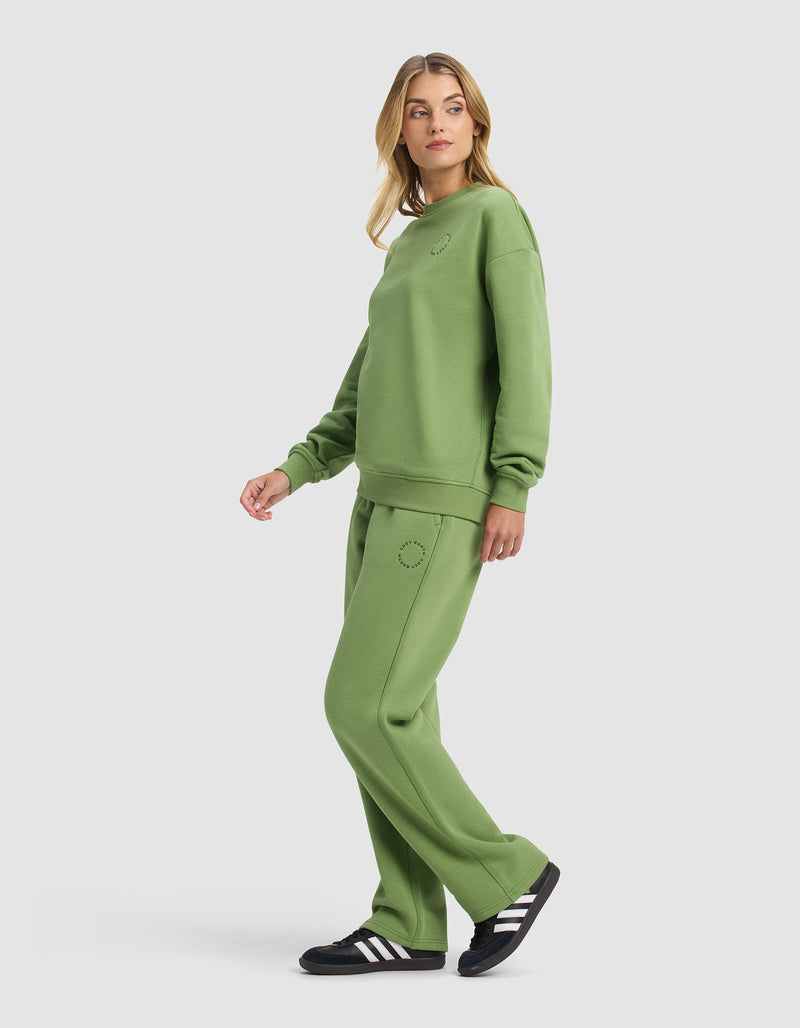 A woman stands sideways wearing the Cozy Earth Women's Cityscape 2.0 Crewneck & Wide Leg Pant Set in green with black and white sneakers. The loose, casual outfit is shown against a plain white background as she looks over her shoulder.