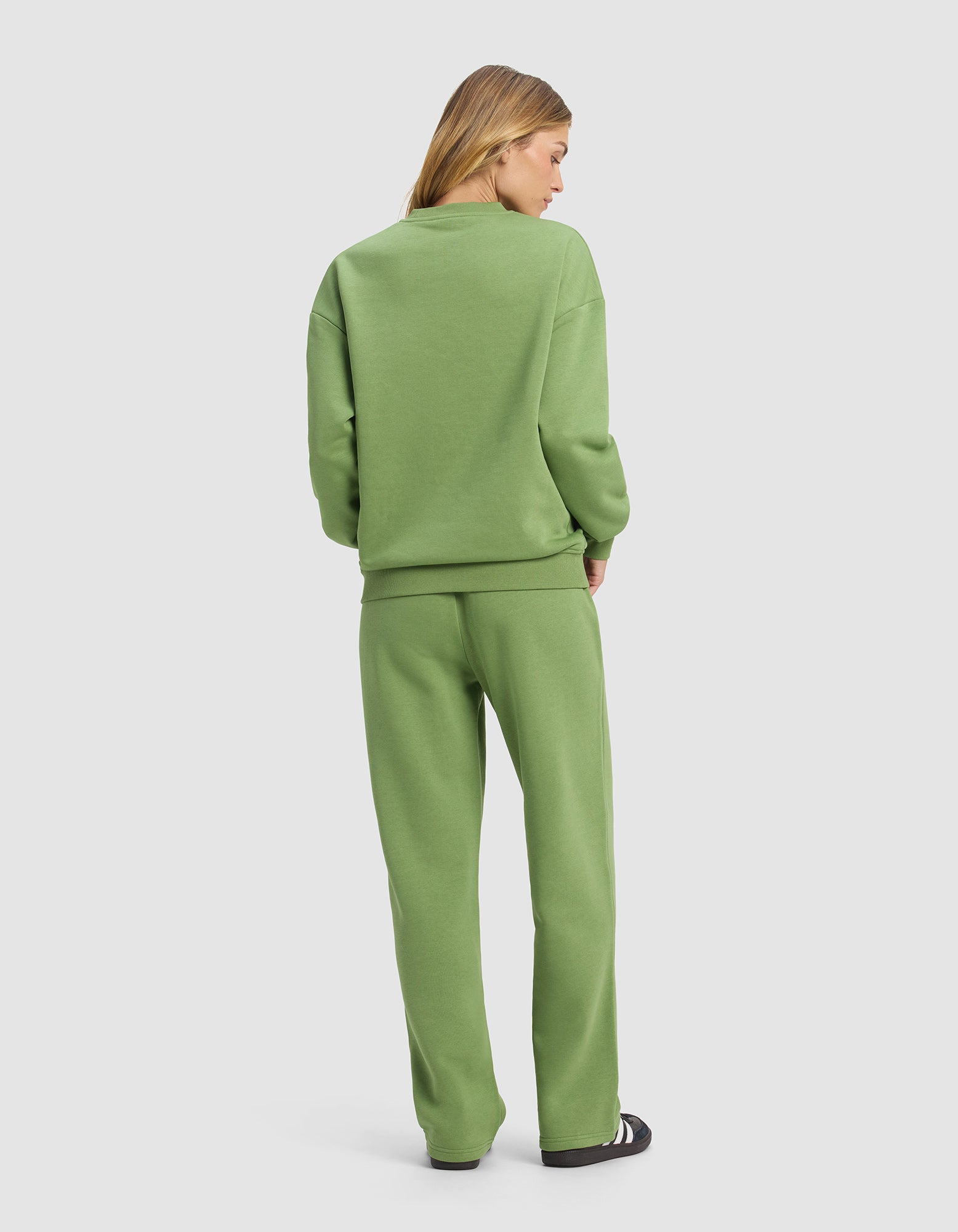 A woman faces away, wearing the Cozy Earth Women's Cityscape 2.0 Crewneck & Wide Leg Pant Set in light green with black and white sneakers, standing against a plain light gray background.|Color:Clover