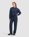 A woman wears the Cozy Earth Women's Cityscape 2.0 Crewneck & Wide Leg Pant Set in navy, paired with black Adidas sneakers. Her hair is in a ponytail and one hand rests in her pocket against a plain light background.|Color:Eclipse