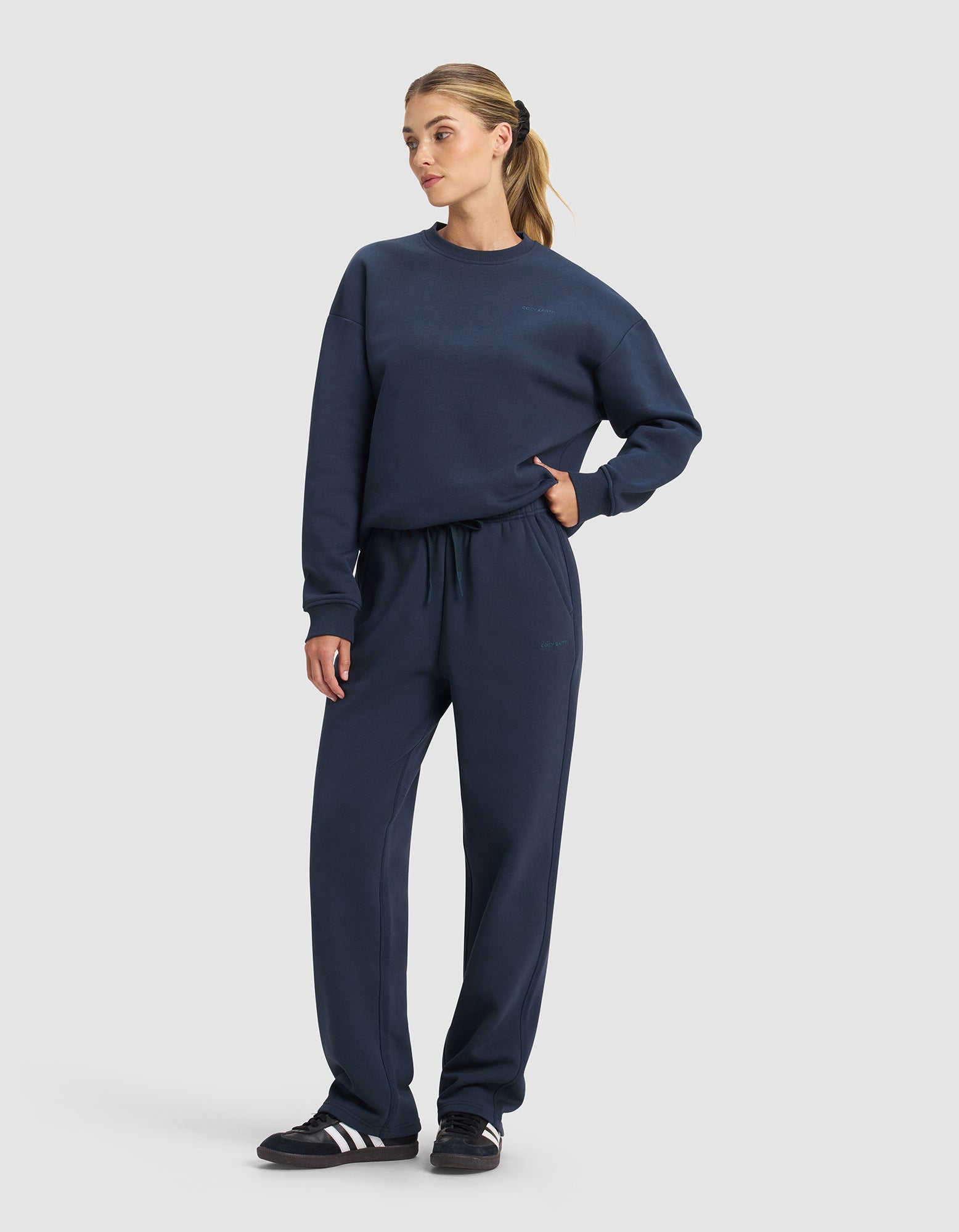A woman wears the Cozy Earth Women's Cityscape 2.0 Crewneck & Wide Leg Pant Set in navy, paired with black Adidas sneakers. Her hair is in a ponytail and one hand rests in her pocket against a plain light background.|Color:Eclipse