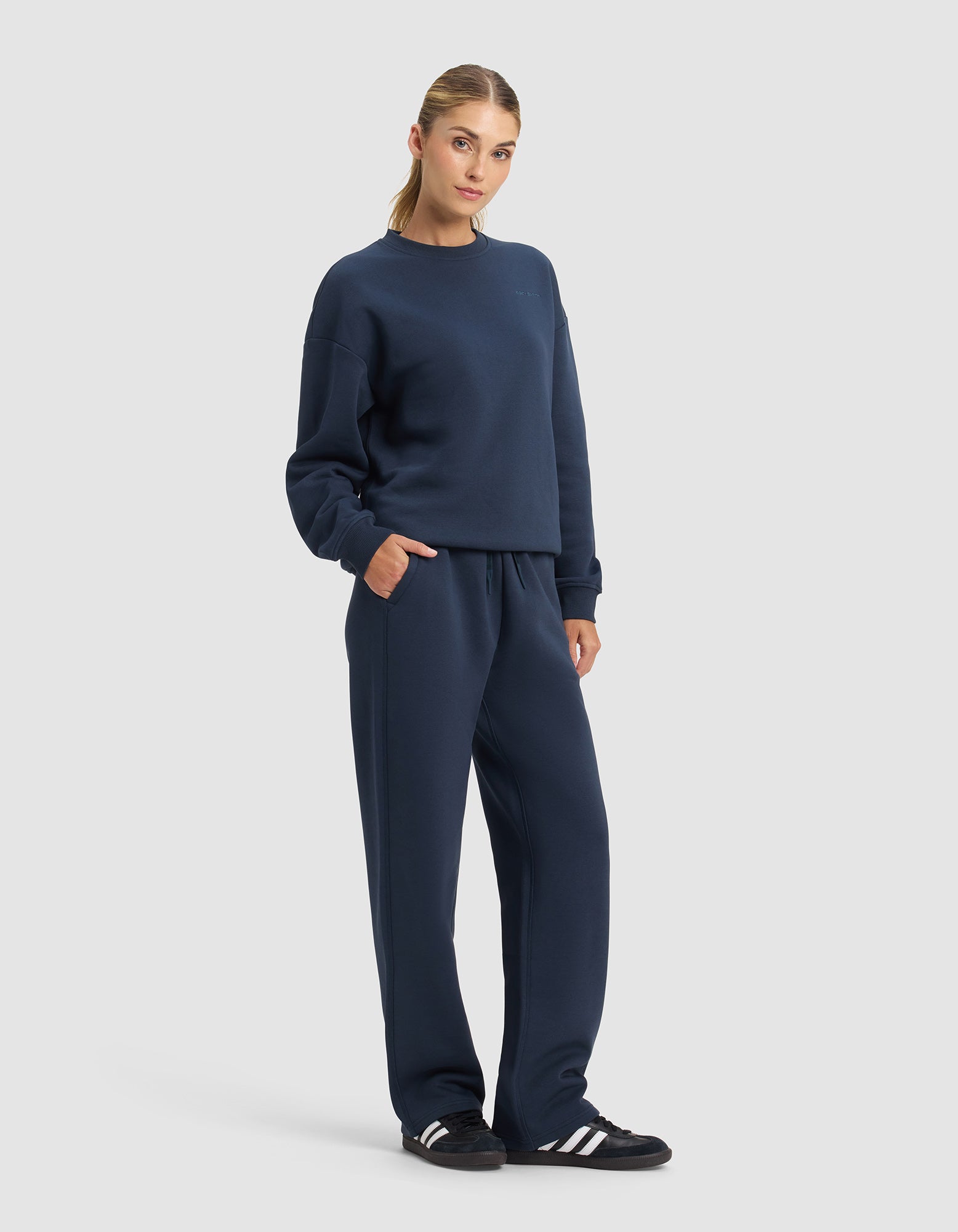 A woman stands against a plain background wearing the Cozy Earth Women's Cityscape 2.0 Crewneck & Wide Leg Pant Set with black and white sneakers, one hand in her pocket, looking directly at the camera.