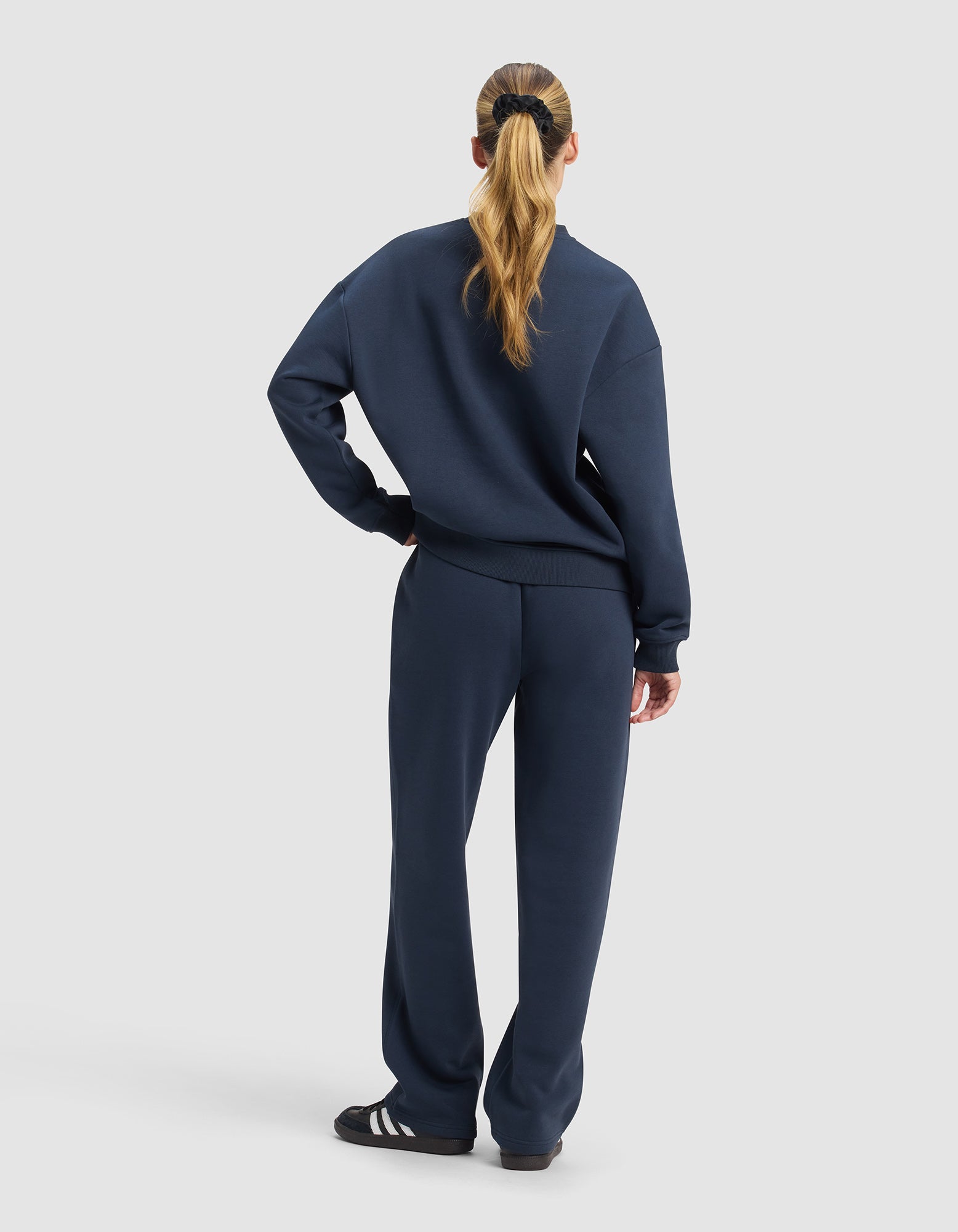 A woman with long hair in a ponytail faces away, wearing the Cozy Earth Women's Cityscape 2.0 Crewneck & Wide Leg Pant Set in navy blue and black sneakers with white stripes. The background is plain light gray.
