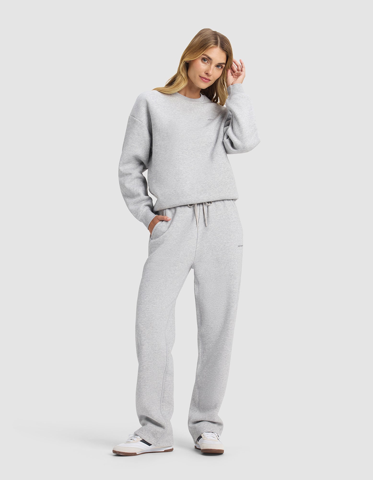 A woman models the Cozy Earth Women's Cityscape 2.0 Crewneck & Wide Leg Pant Set in light gray, paired with white sneakers. She stands against a plain background, one hand in her pocket and the other touching her hair, gazing calmly at the camera.