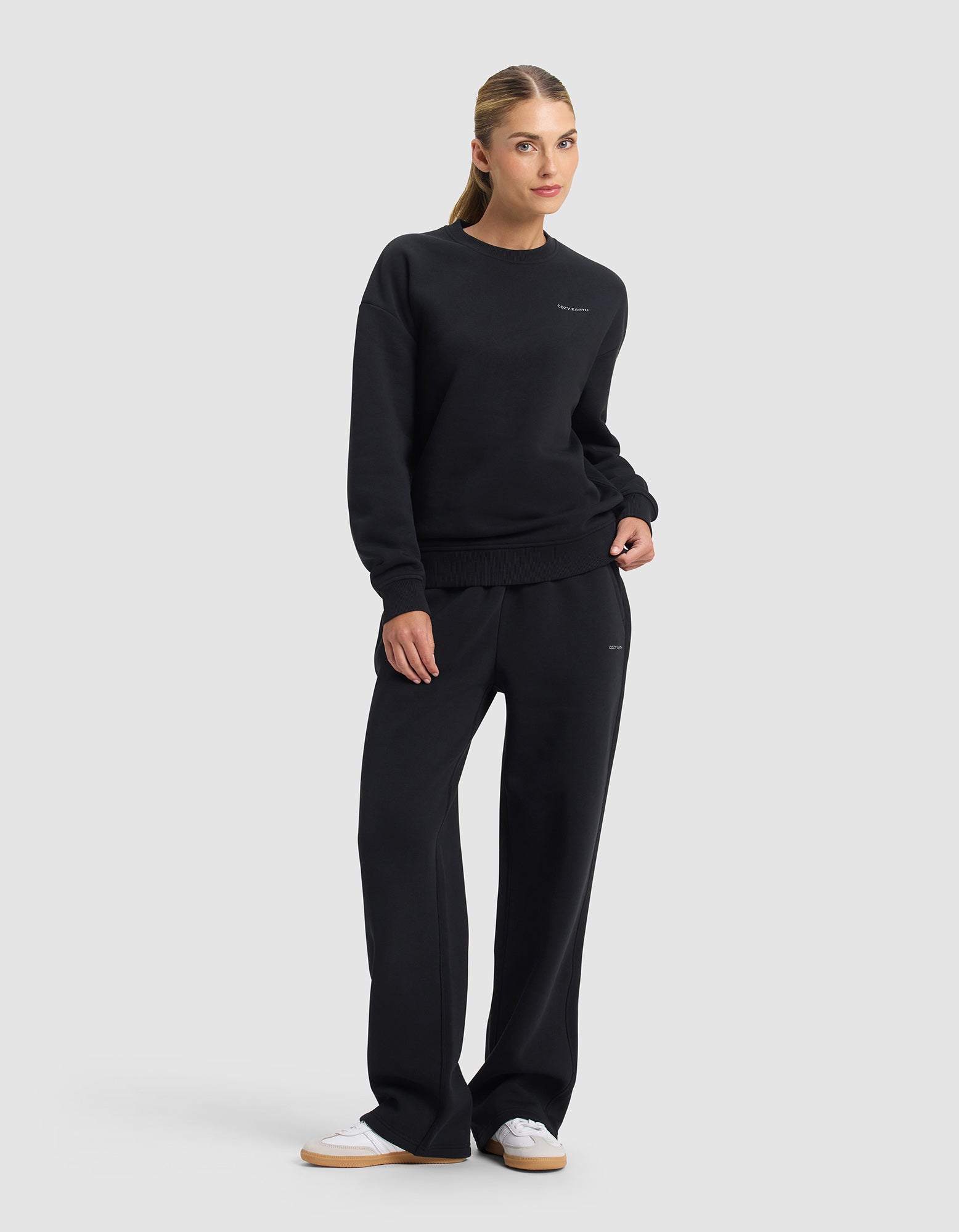 A woman with light brown hair pulled back models the Cozy Earth Women's Cityscape 2.0 Crewneck & Wide Leg Pant Set in black, paired with white sneakers, standing against a plain background with one hand in her pocket.|Color:Jet Black