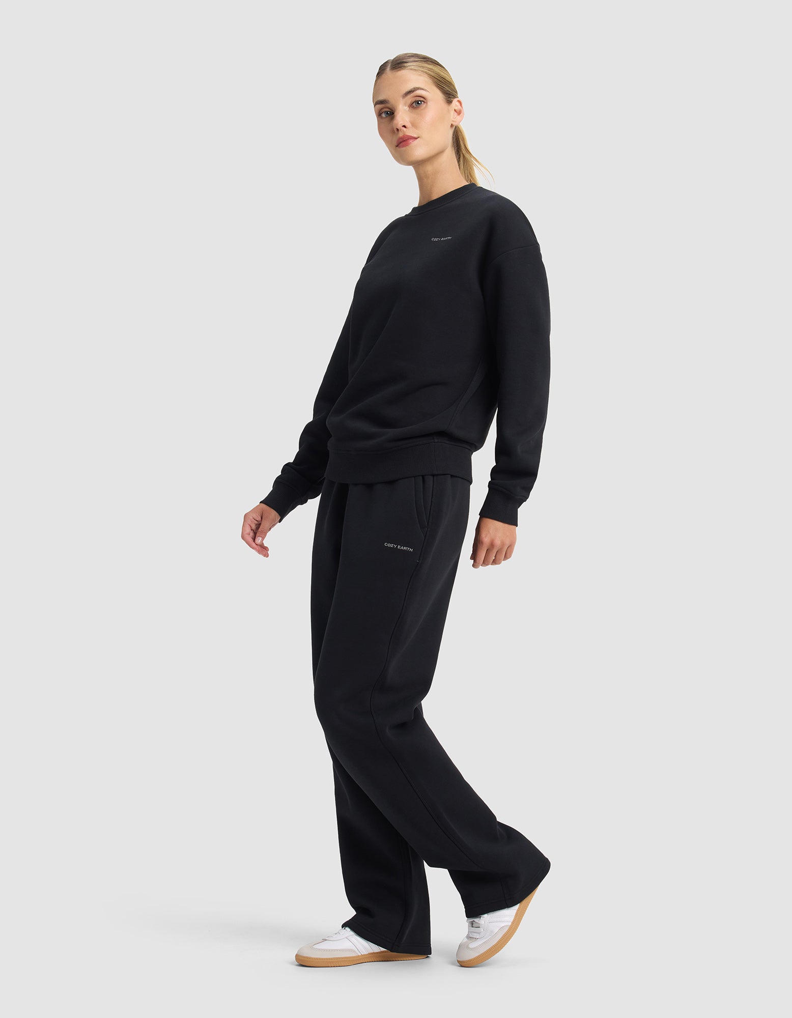 A woman models the Cozy Earth Women's Cityscape 2.0 Crewneck & Wide Leg Pant Set in black with white sneakers, standing against a plain light background, looking to the side with relaxed arms and one foot forward.|Color:Jet Black