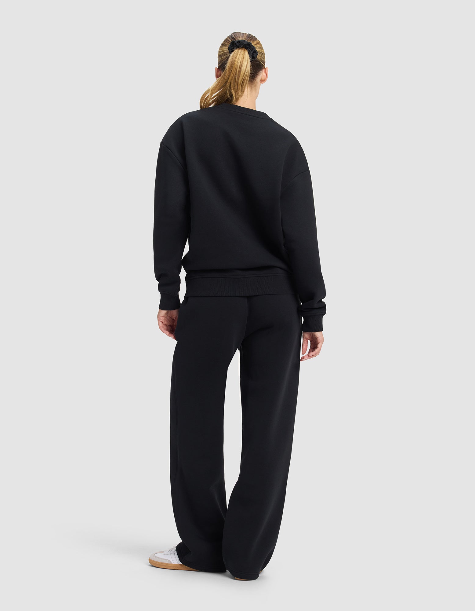 A woman with a ponytail faces away, wearing the Cozy Earth Women's Cityscape 2.0 Crewneck & Wide Leg Pant Set in black with white sneakers, set against a plain light gray background.|Color:Jet Black
