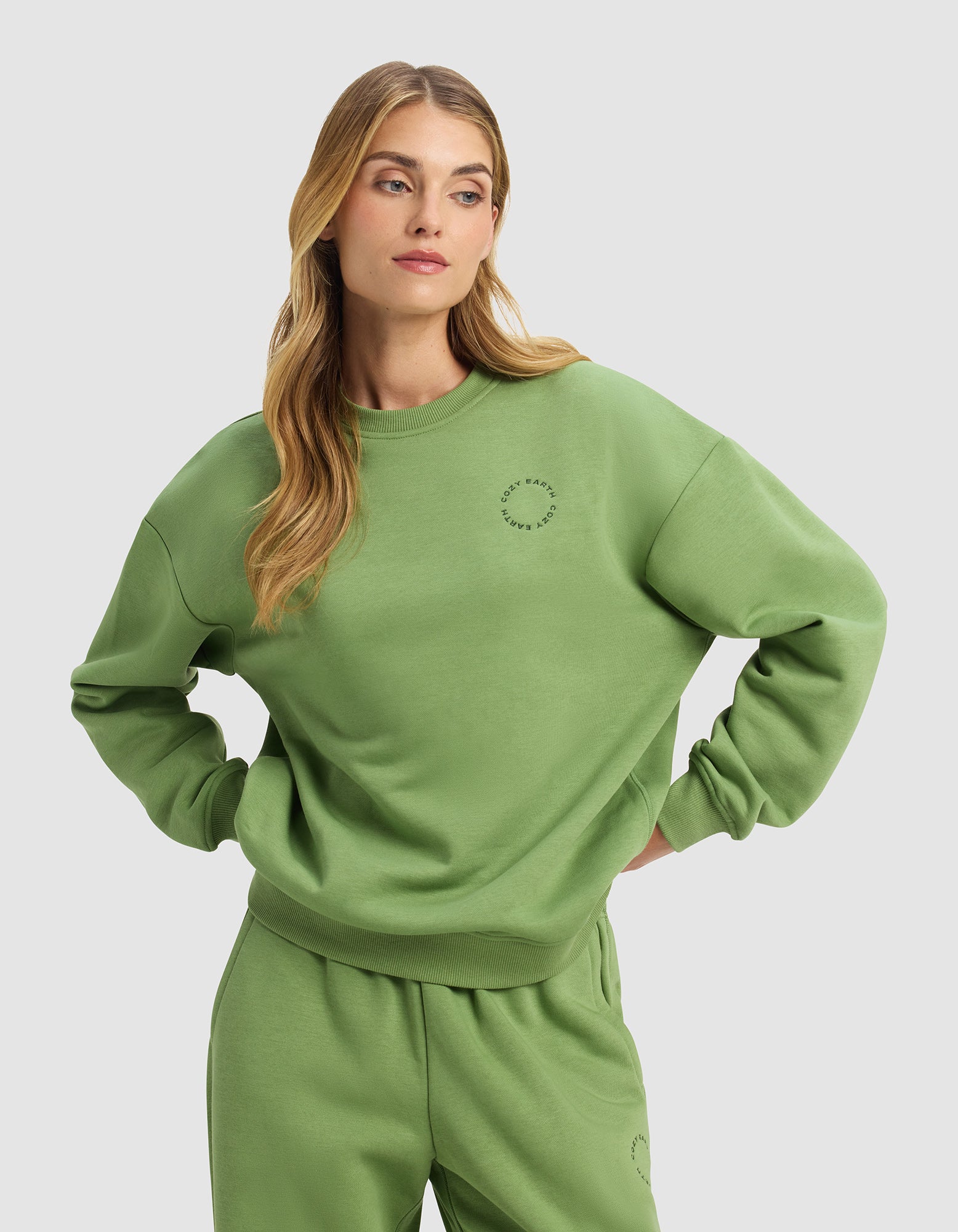 A woman with long blonde hair stands with her hands on her hips against a plain light gray background, wearing the Cozy Earth Women's Cityscape 2.0 Crewneck in a loose-fitting, light green style with matching sweatpants.