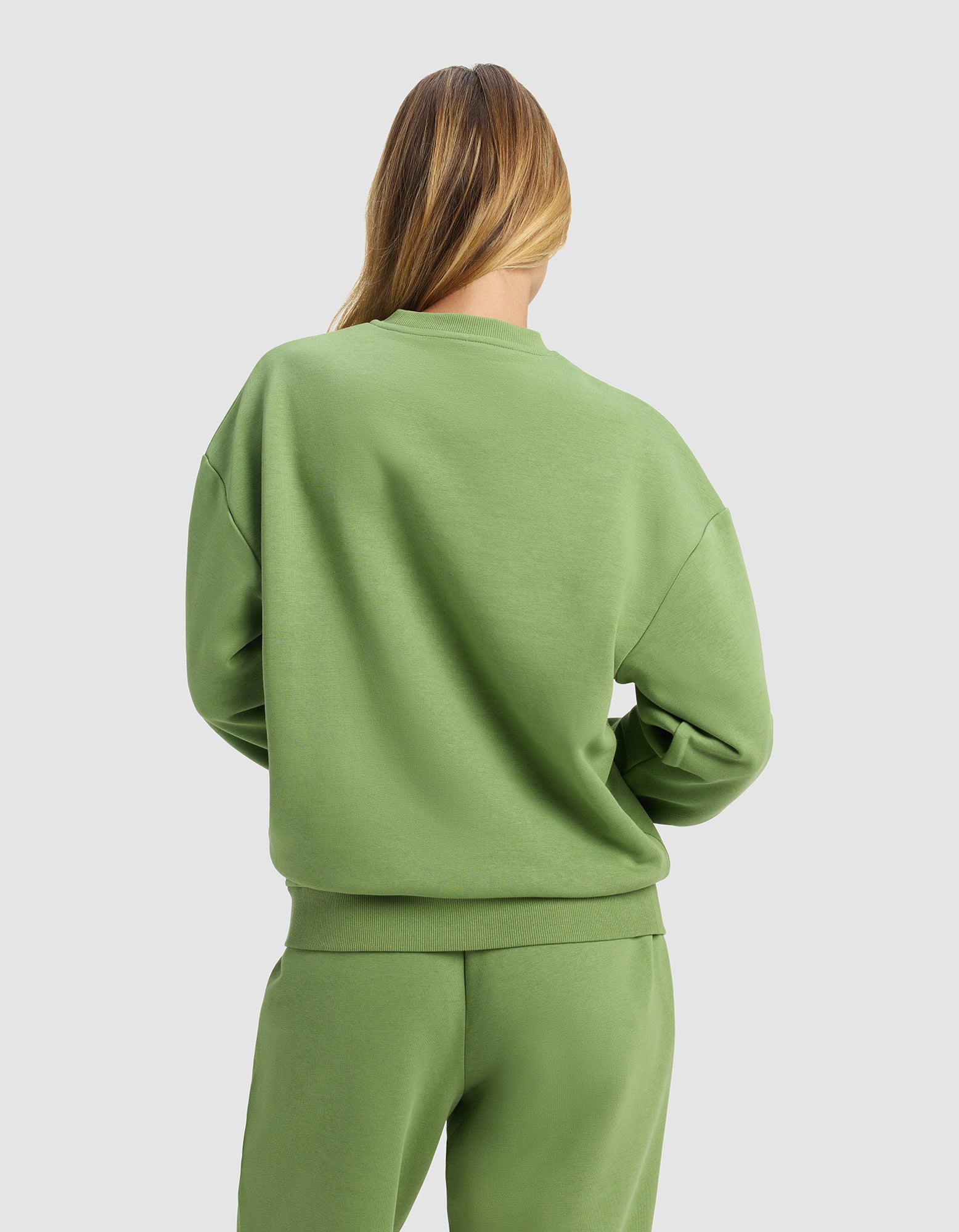A woman with long, light brown hair is seen from behind wearing the Cozy Earth Women's Cityscape 2.0 Crewneck in light green, paired with matching sweatpants, standing against a plain light gray background.|Color:Clover