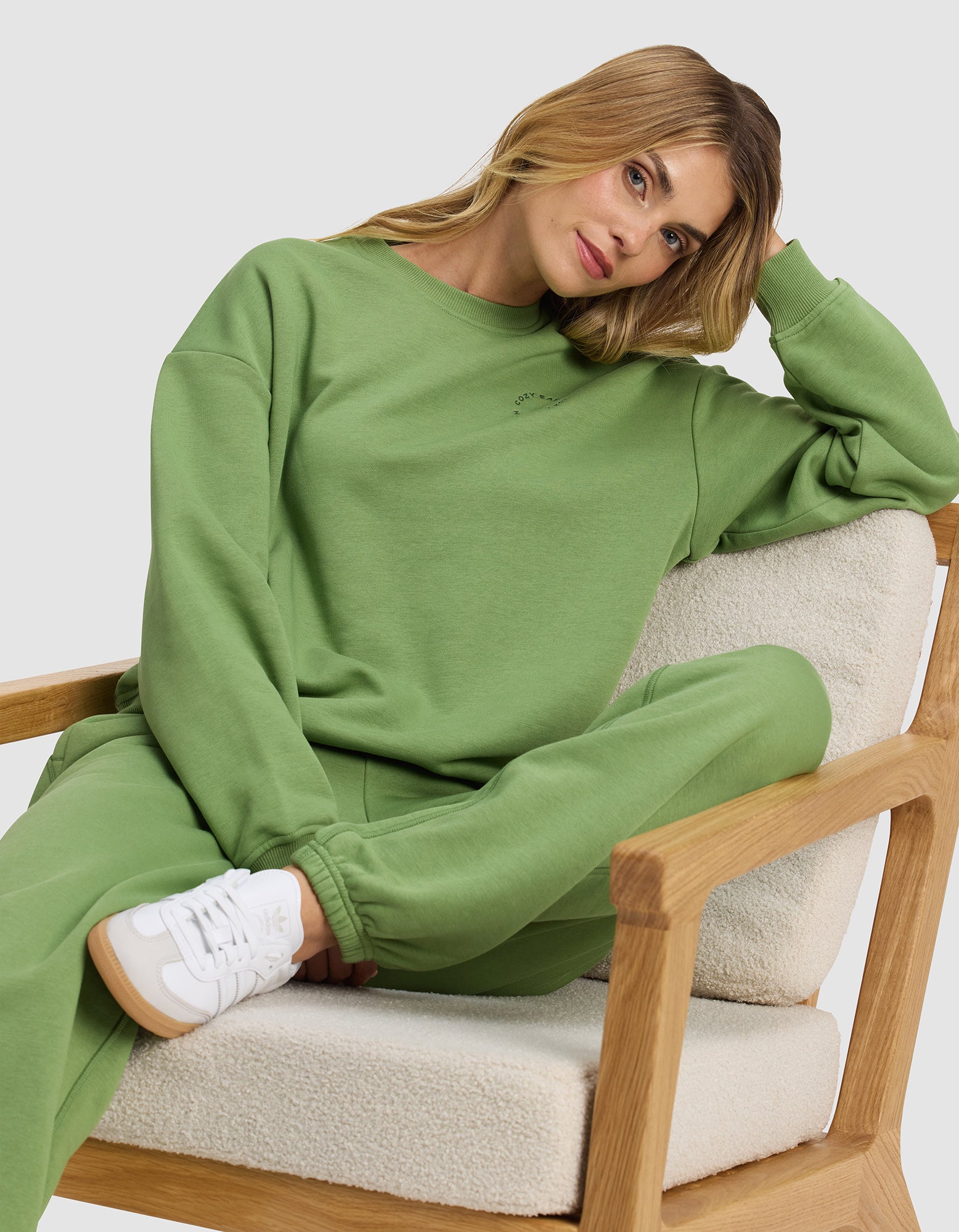 A woman wears the Cozy Earth Women's Cityscape 2.0 Crewneck, lounging casually on a cushioned wooden chair and looking relaxed in her stylish, comfortable outfit.|Color:Clover