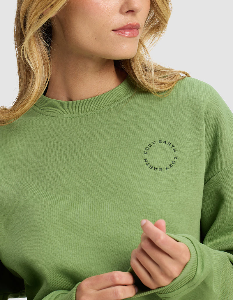 A woman wearing the Cozy Earth Women's Cityscape 2.0 Crewneck in light green poses against a plain background, facing slightly to the side with her hand near her collar.