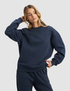 A woman with long blonde hair models the Cozy Earth Women's Cityscape 2.0 Crewneck in navy, paired with matching sweatpants. She stands with one hand behind her head and the other on her hip, gazing off to the side.|Color:Eclipse